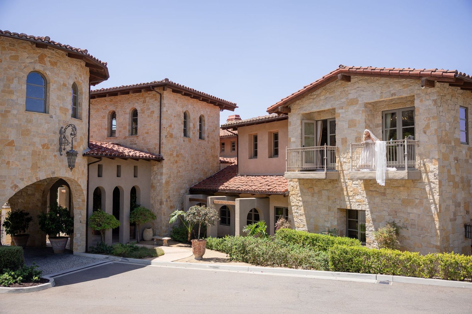 Elegant Mediterranean-style stone villa with red tile roof, arched walkways, and balcony under bright blue sky.