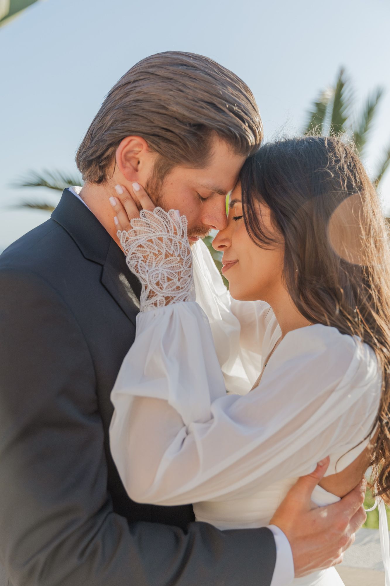 A bride and groom share an intimate forehead-to-forehead moment outdoors, with the bride wearing elegant lace-sleeved white gown.