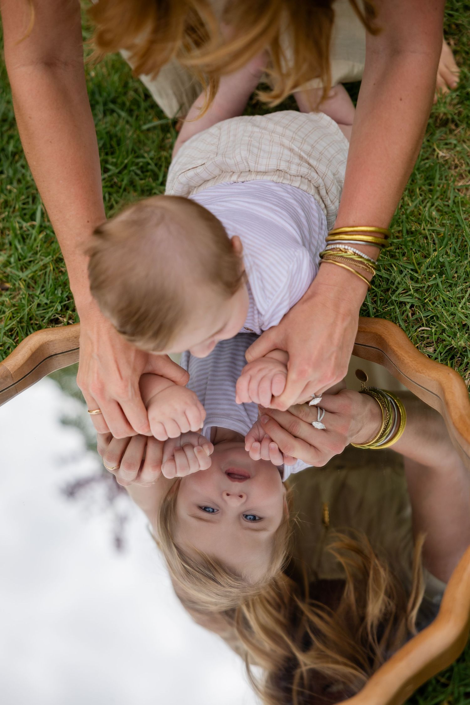 Mother holding baby over mirror on grass, reflecting blonde woman's face with gold bangles and engagement ring.