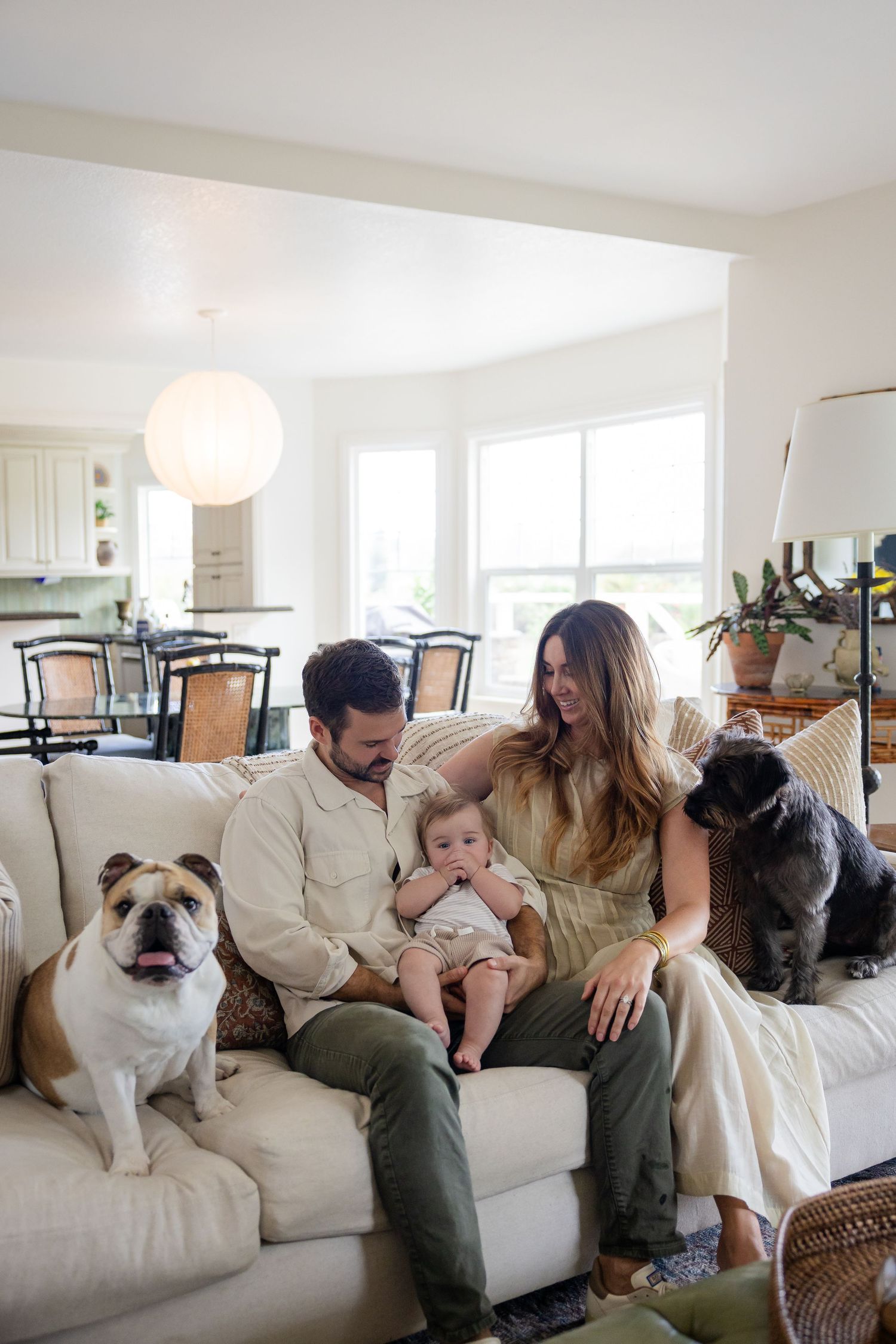 Happy couple sitting on white sofa with baby and two dogs in bright modern living room.