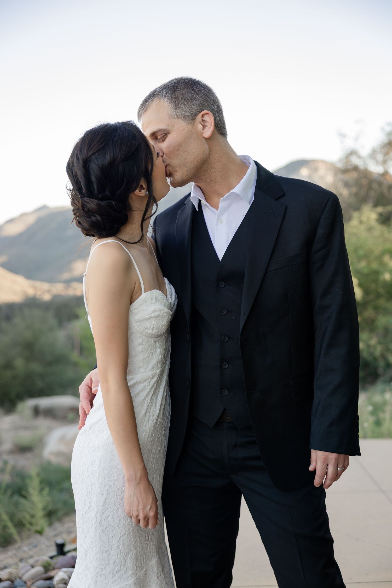 Bride and groom share a tender kiss outdoors with scenic mountain landscape in the background at dusk.