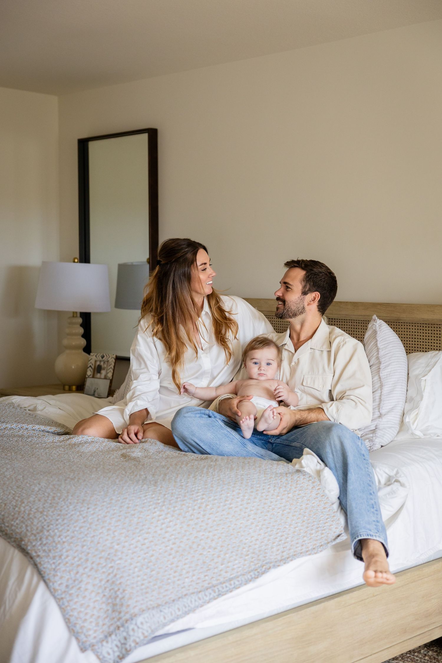 Happy young family with baby sitting together on a bed in a bright, cozy bedroom.