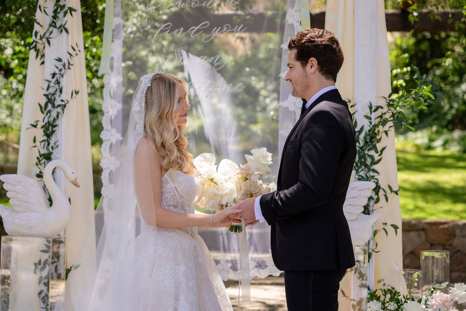 Bride and groom exchange vows at an elegant outdoor wedding ceremony with white floral decor and lush greenery.