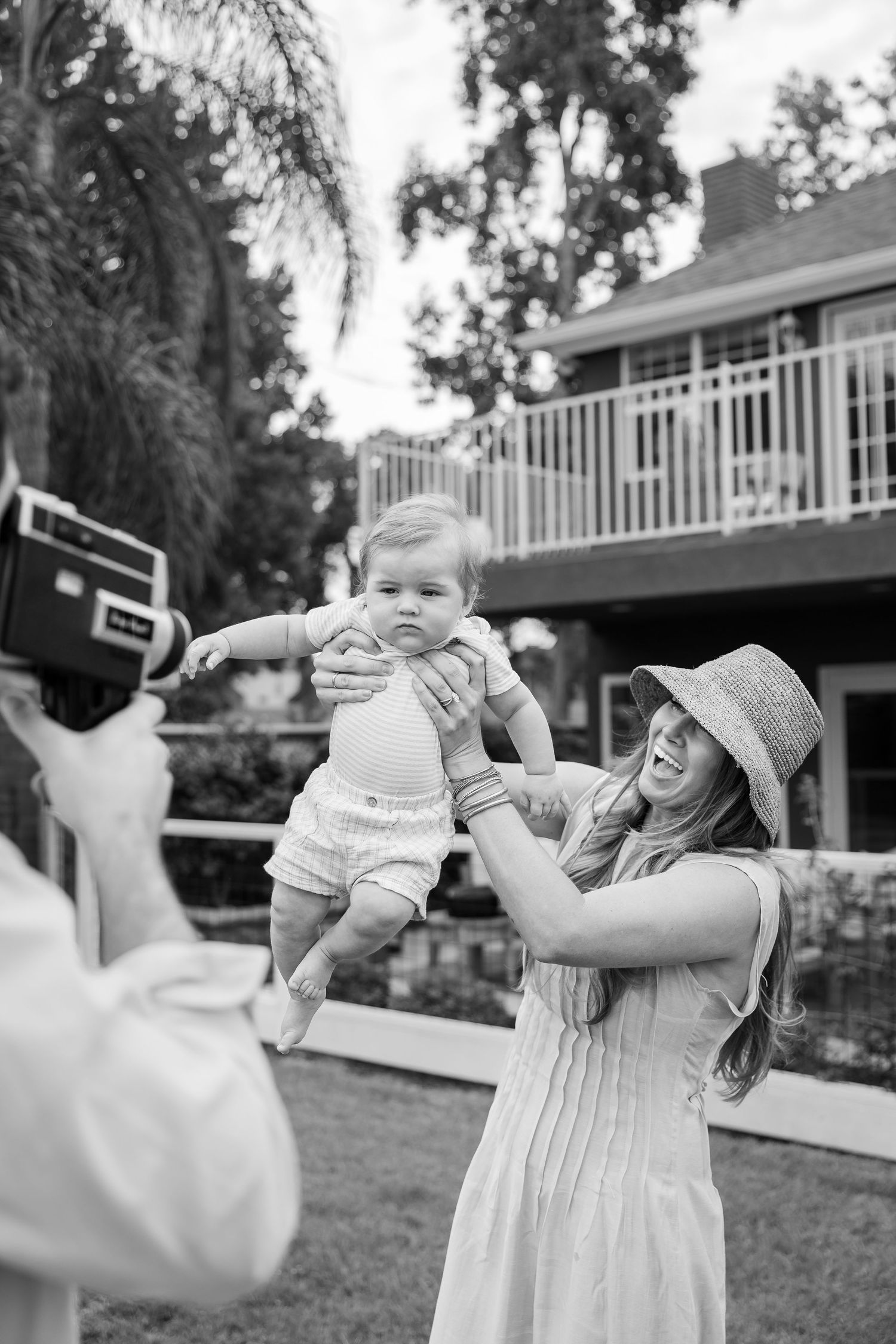 Black and white photo of a smiling woman in a sun hat lifting a baby outdoors while someone films with a vintage camera.