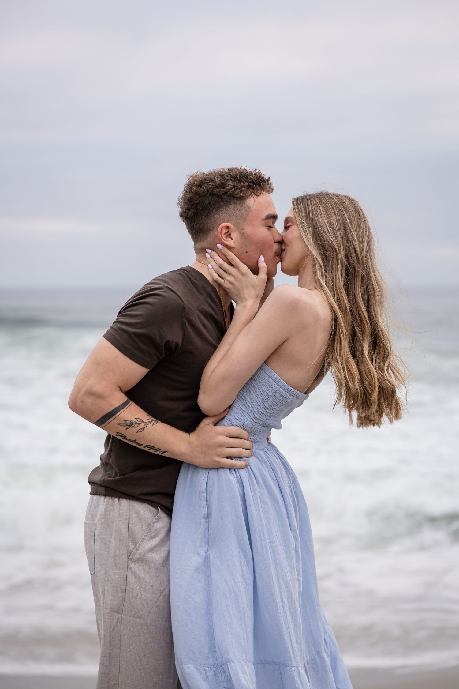 Couple sharing a romantic kiss on the beach, woman in blue dress, man in brown shirt, ocean waves in background.