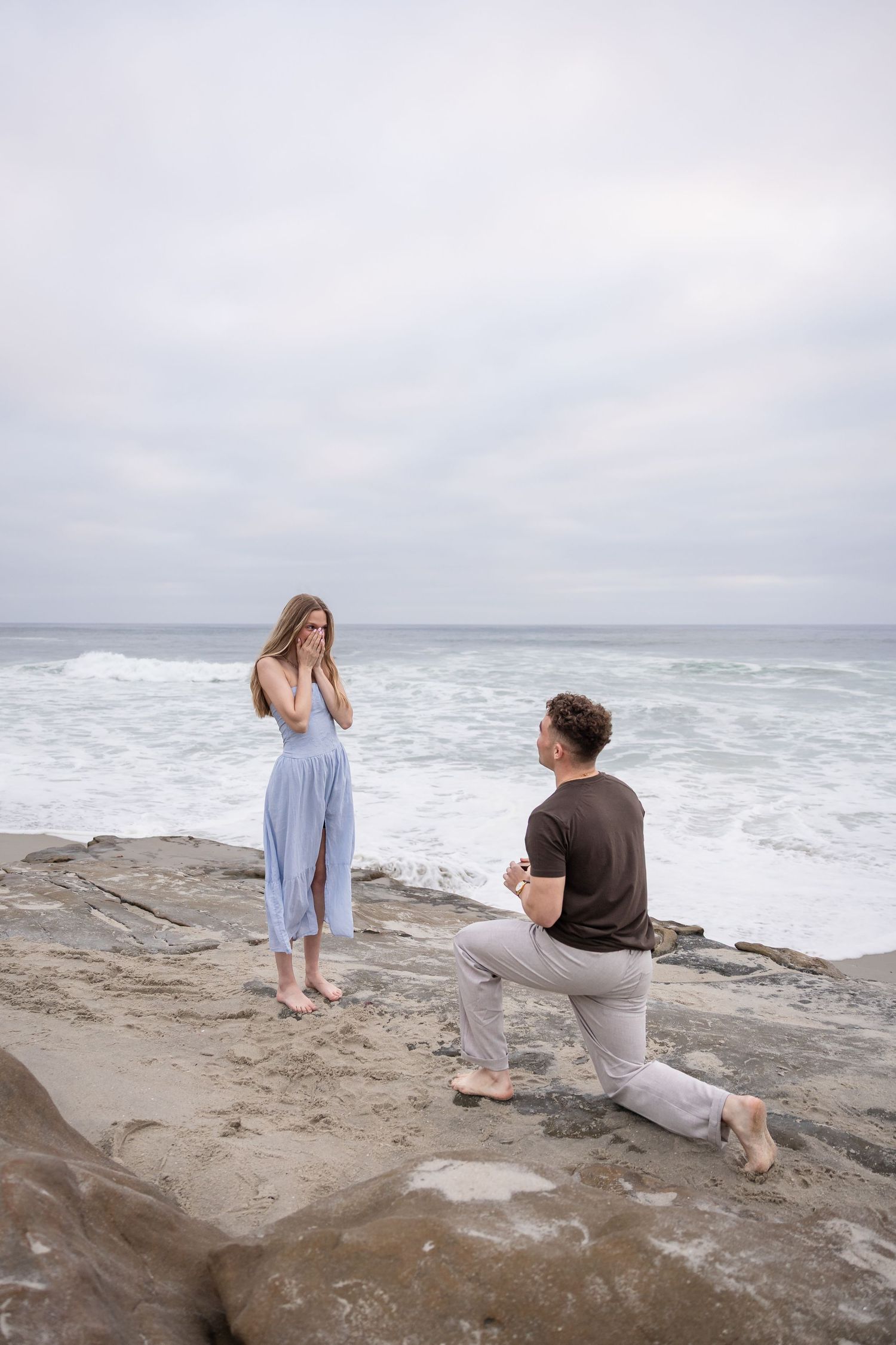 Man proposes on one knee to woman on rocky beach with ocean waves, as she covers her mouth in surprise.