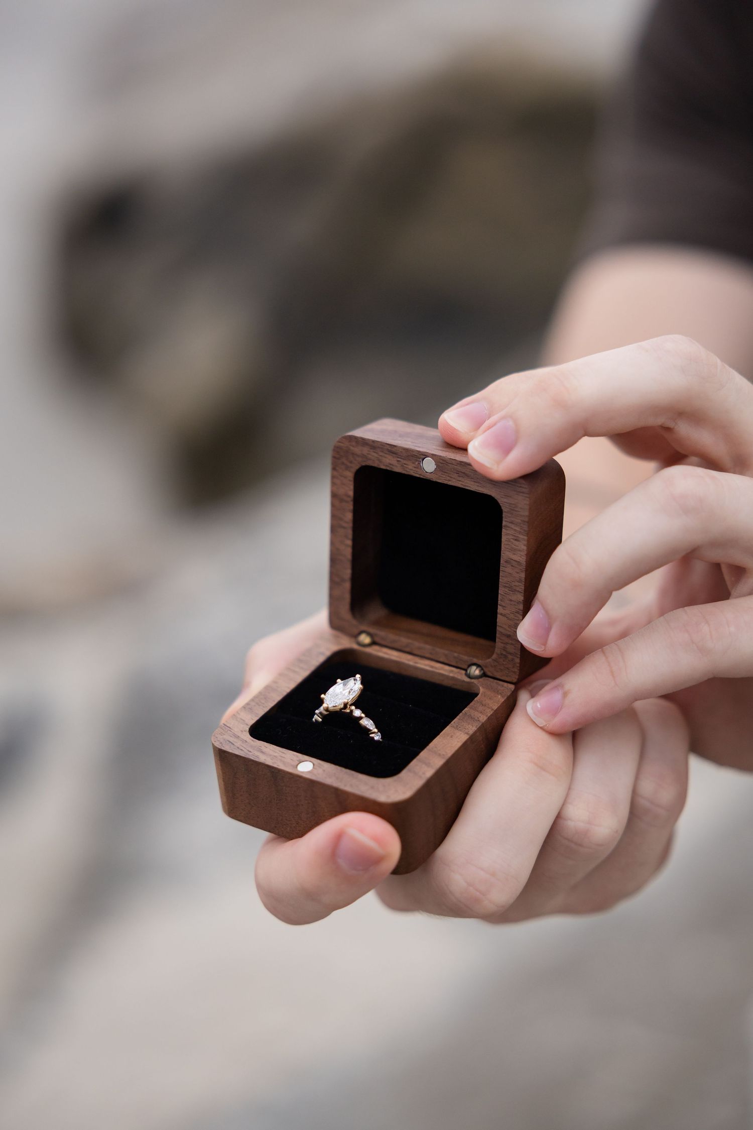 Hands holding open a wooden ring box with a diamond engagement ring on black velvet lining.