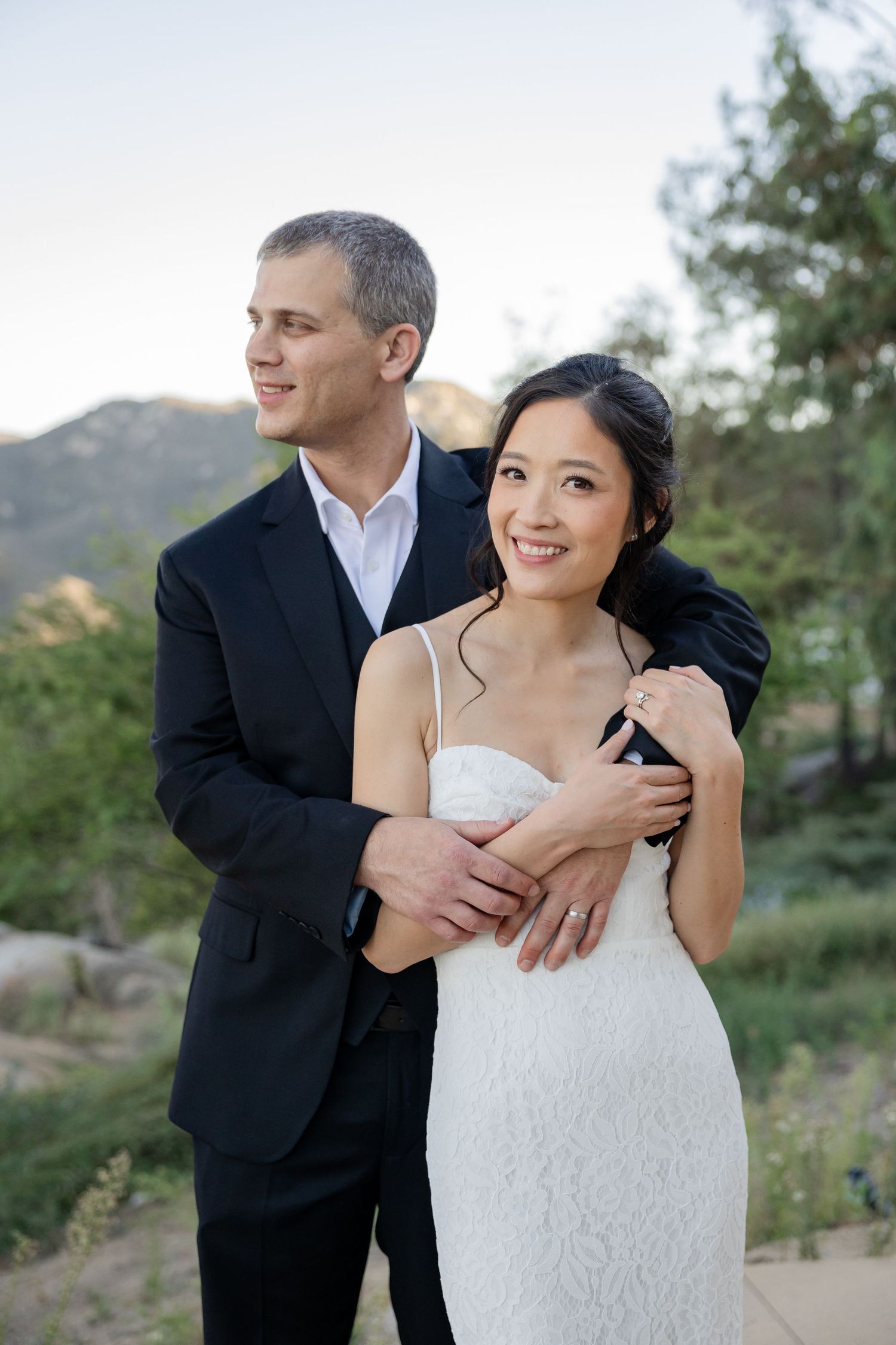 Groom embraces bride from behind as they smile together with a scenic mountain backdrop at sunset.