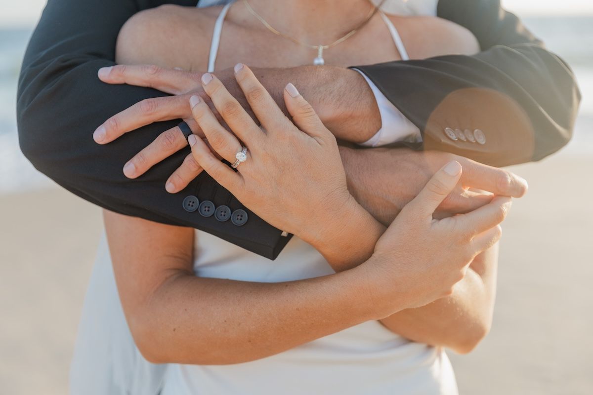 Couple embracing on beach, woman displaying diamond engagement ring on hand wrapped around man in suit jacket.