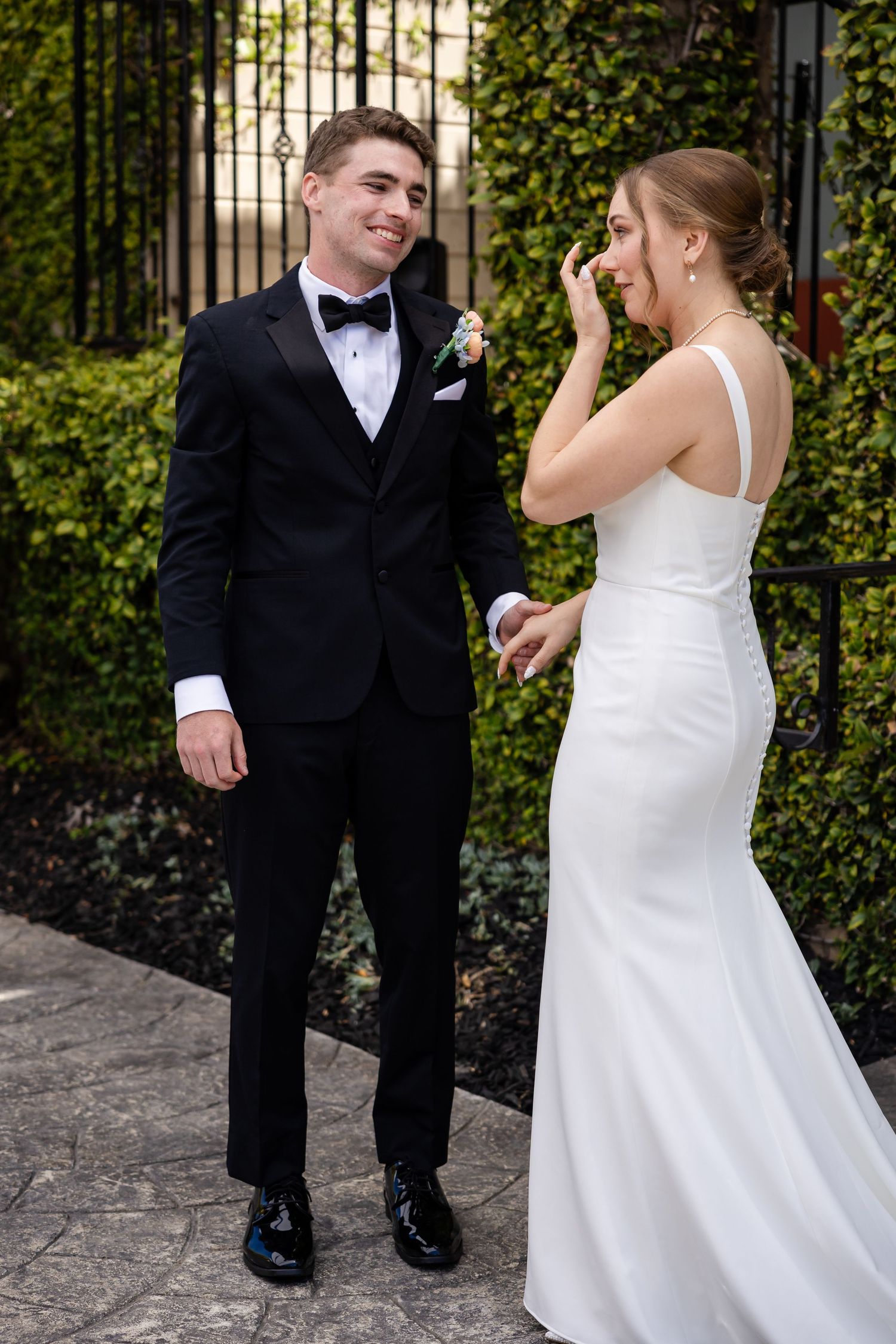Emotional bride in white gown wipes tears during first look with smiling groom in black tuxedo.