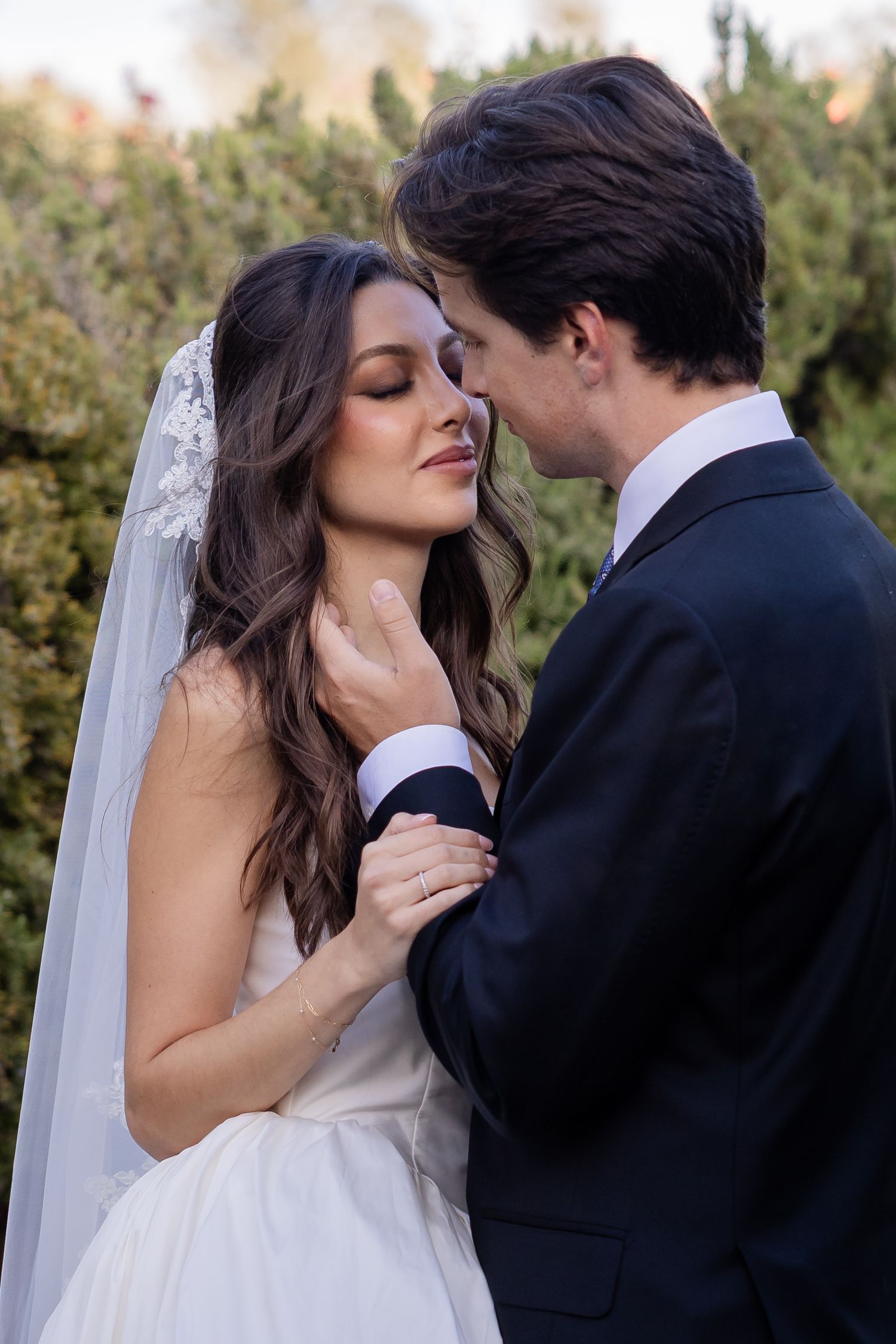 A bride and groom share an intimate moment outdoors, with the groom gently holding the bride's neck as she closes her eyes.