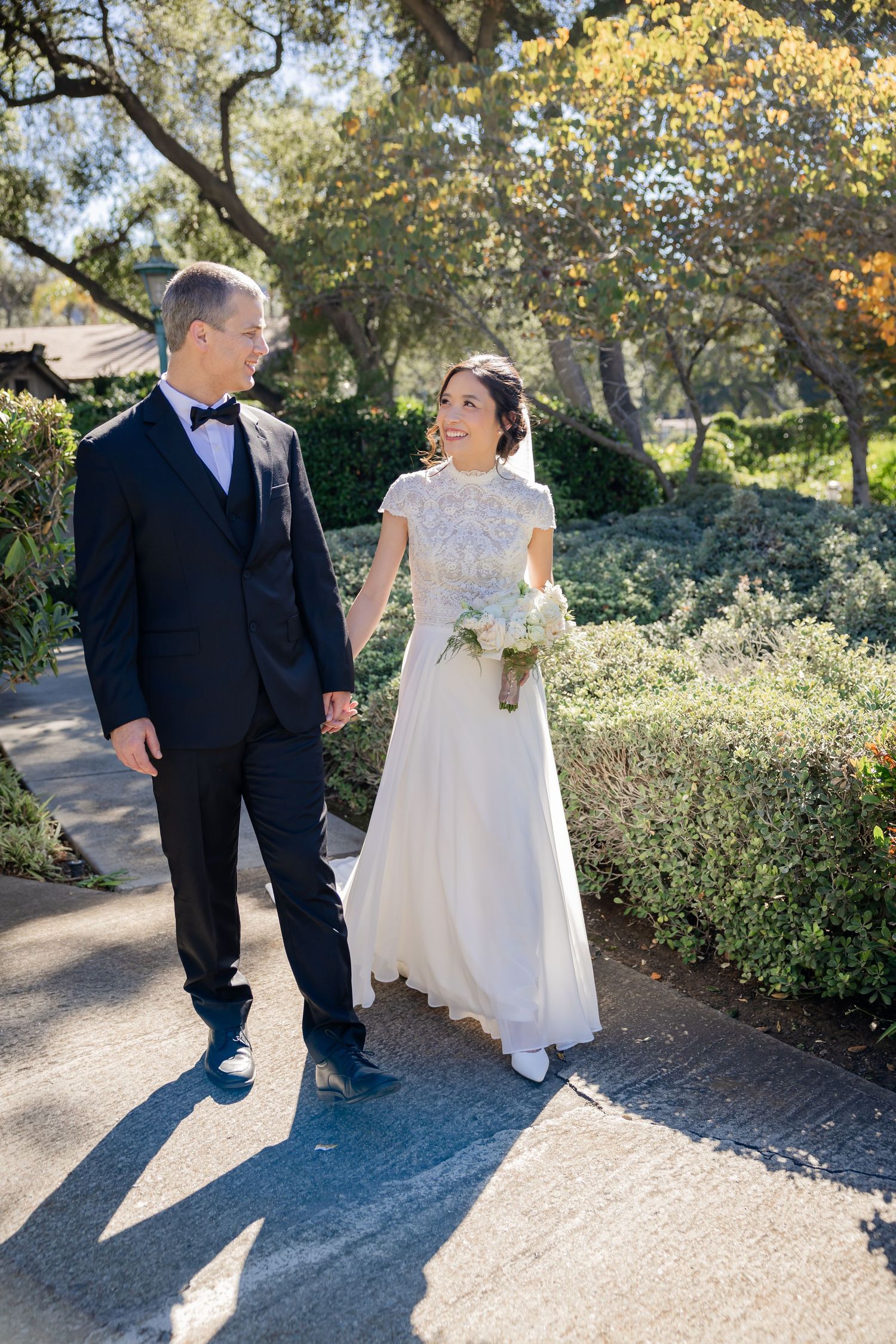 Bride and groom walk hand in hand along a garden path, smiling at each other on their wedding day.