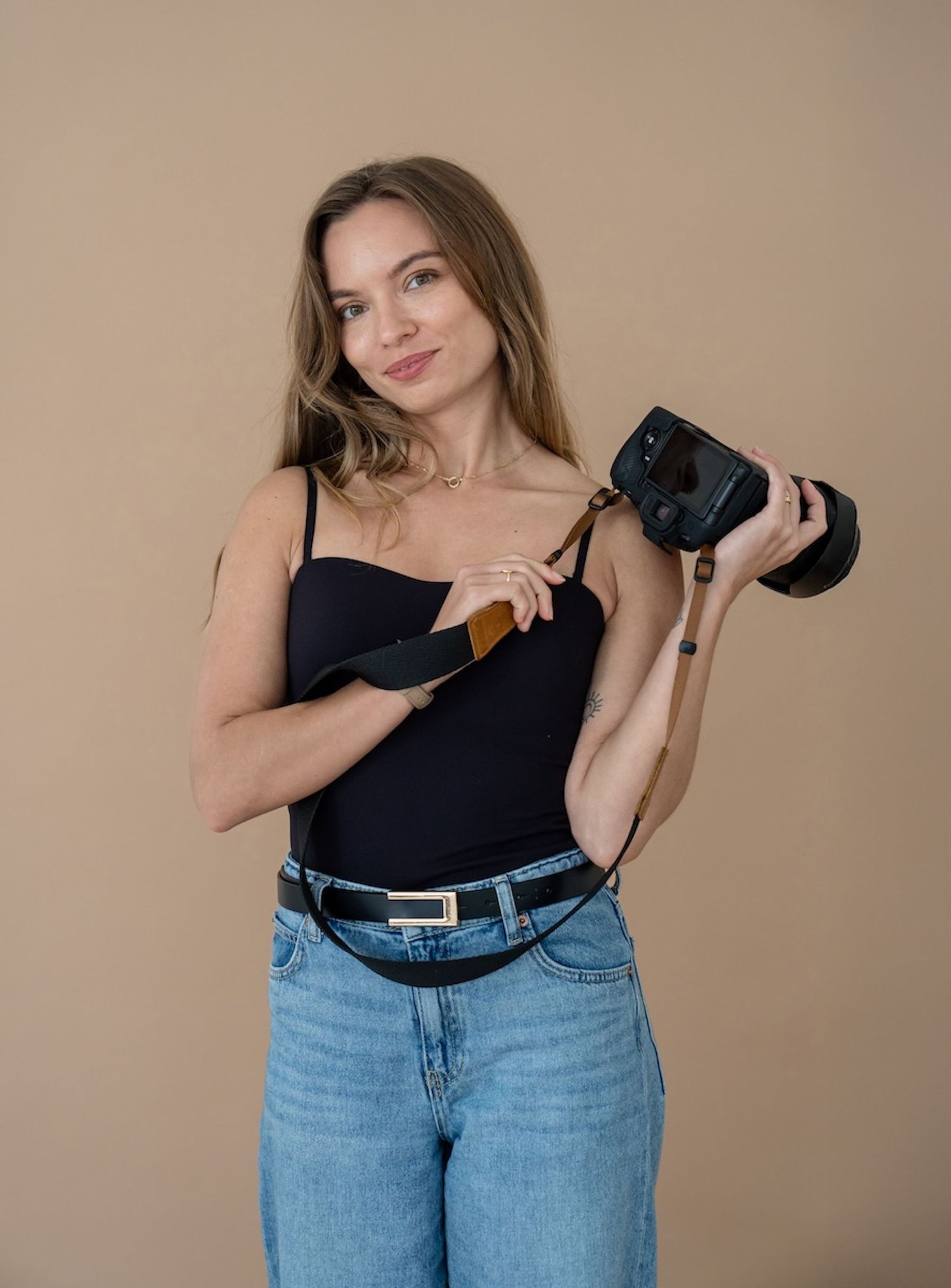 Young woman in black tank top and jeans holding a camera, posing against a beige background.