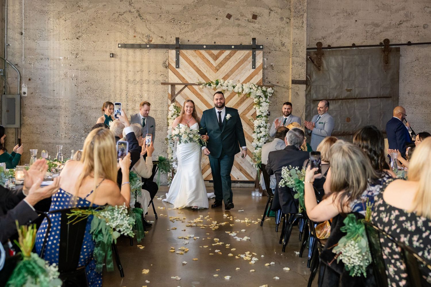 Bride and groom walk down the aisle at an industrial wedding venue with floral arch backdrop.