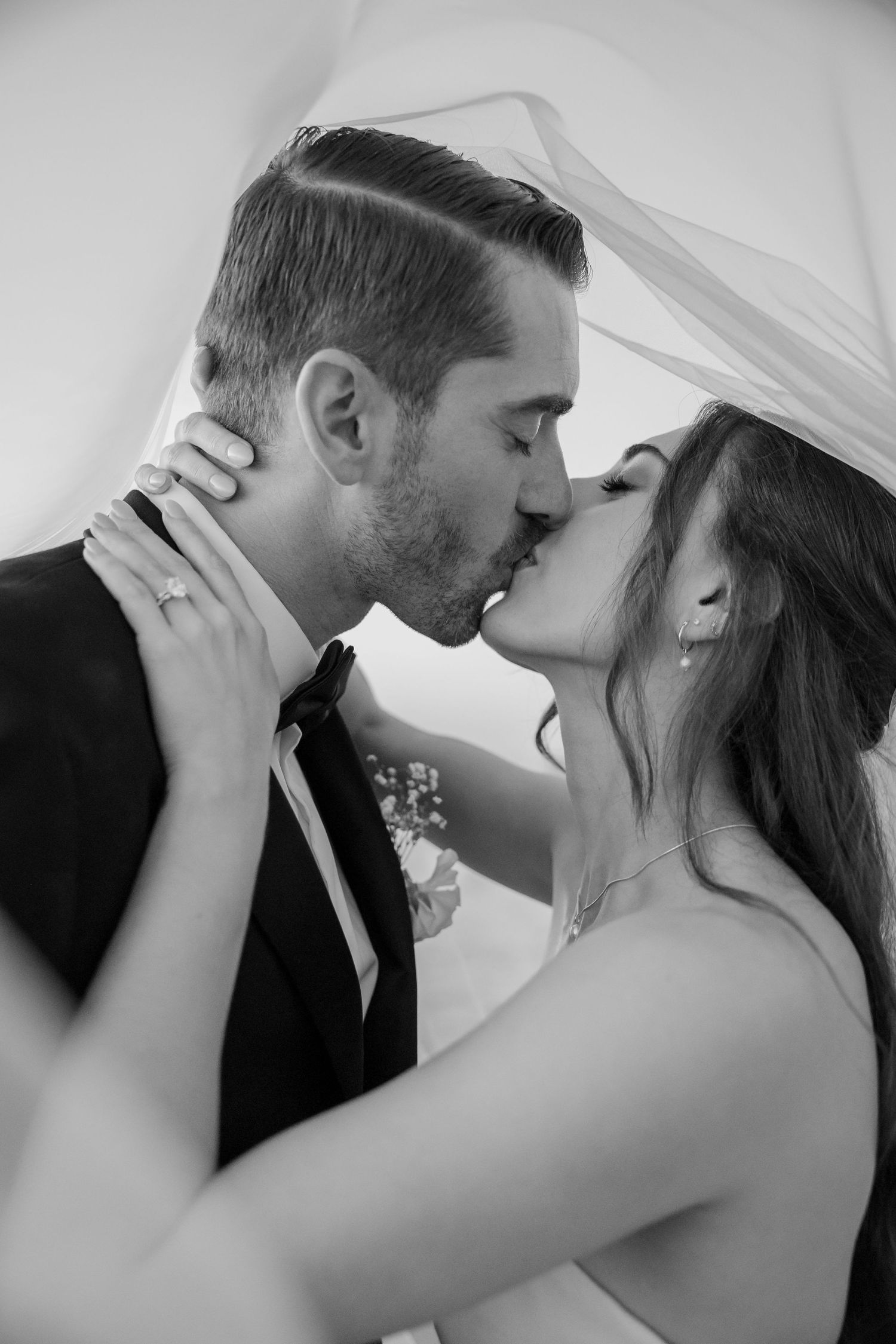 Black and white photo of a bride and groom sharing a romantic kiss under a wedding veil.