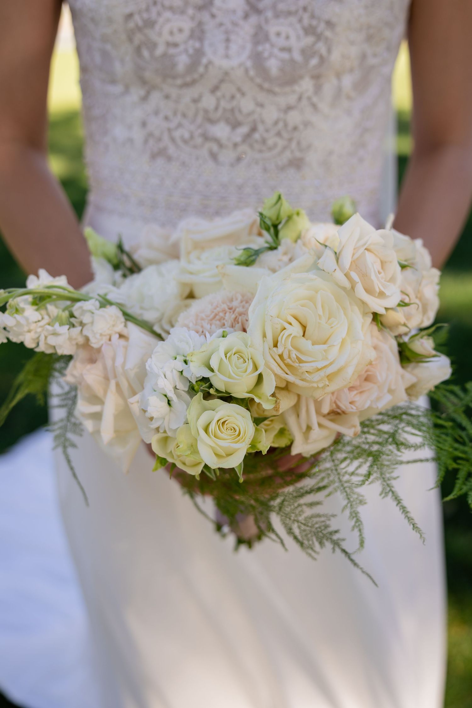 Bride holding elegant white and blush rose wedding bouquet with greenery accents, wearing lace gown.