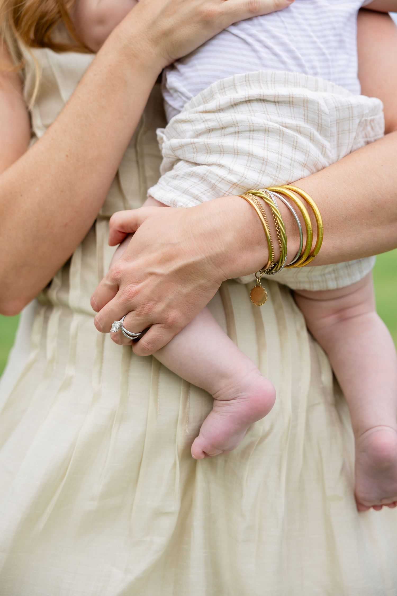 Mother holding baby with gold bangles and silver ring on her wrist, wearing a cream linen dress outdoors.
