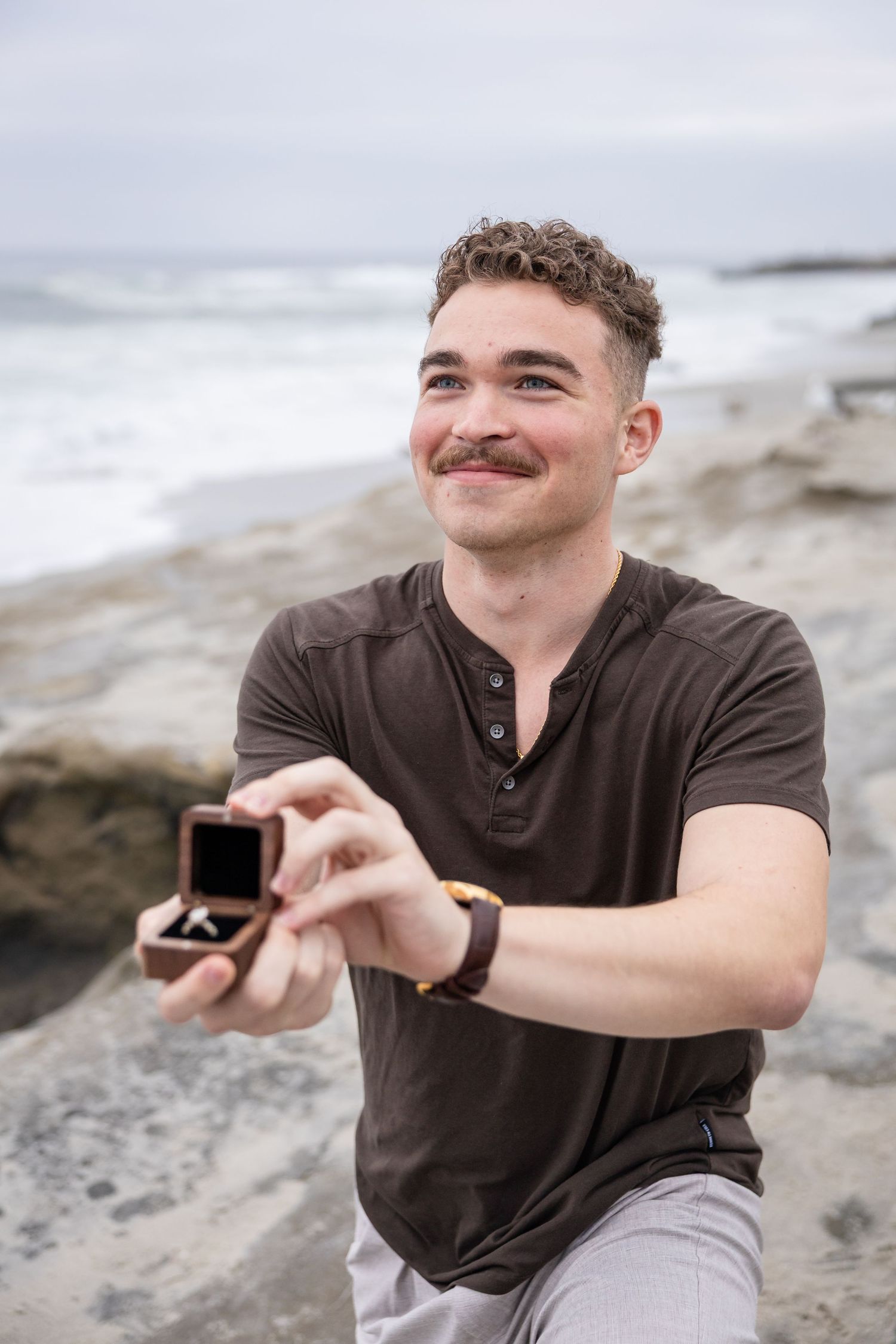Young man with mustache kneeling on rocky beach, smiling while presenting an open ring box for a marriage proposal.