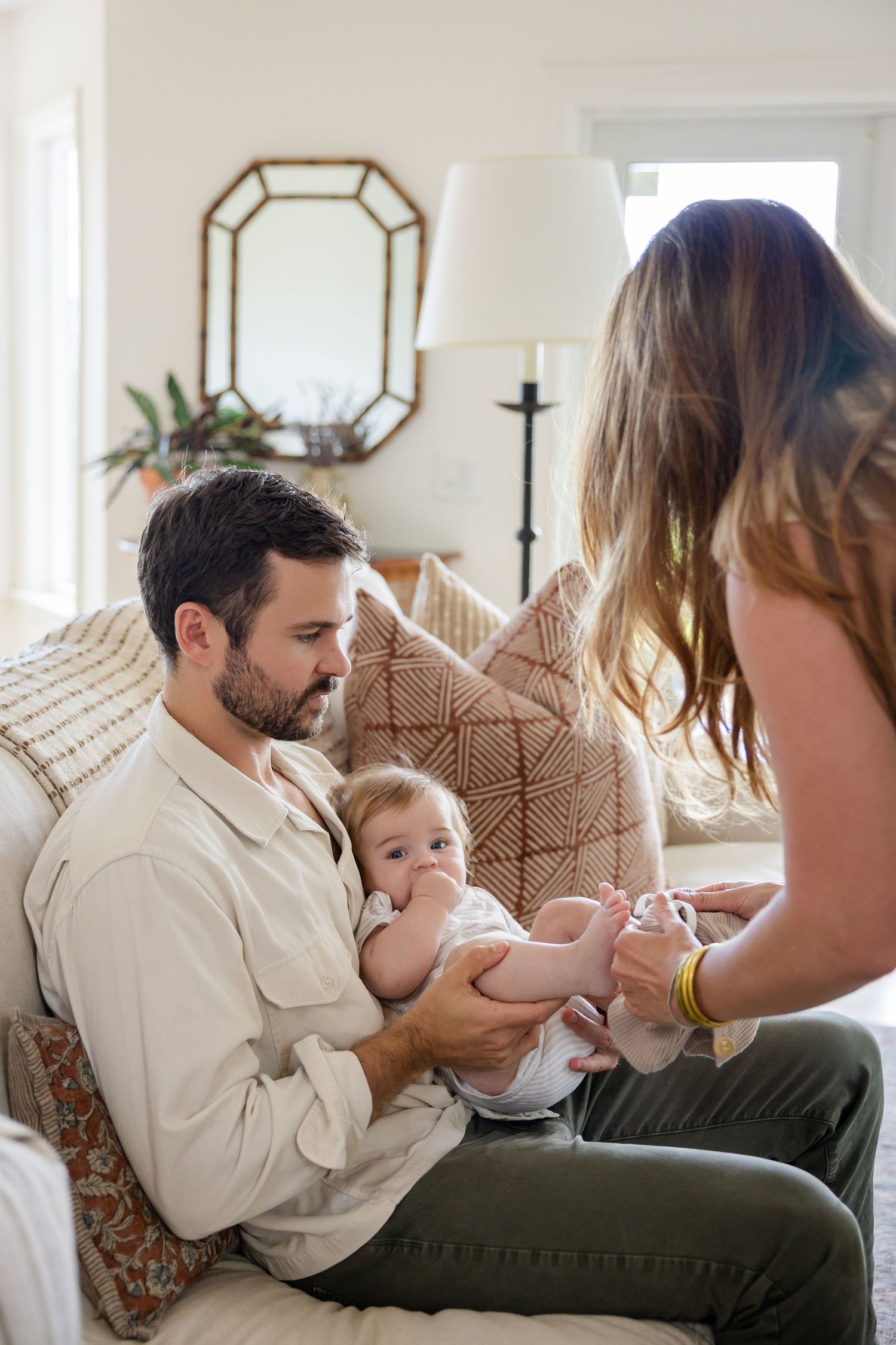 A bearded father holds his baby on a couch while the mother reaches out to interact with the infant in a cozy living room.