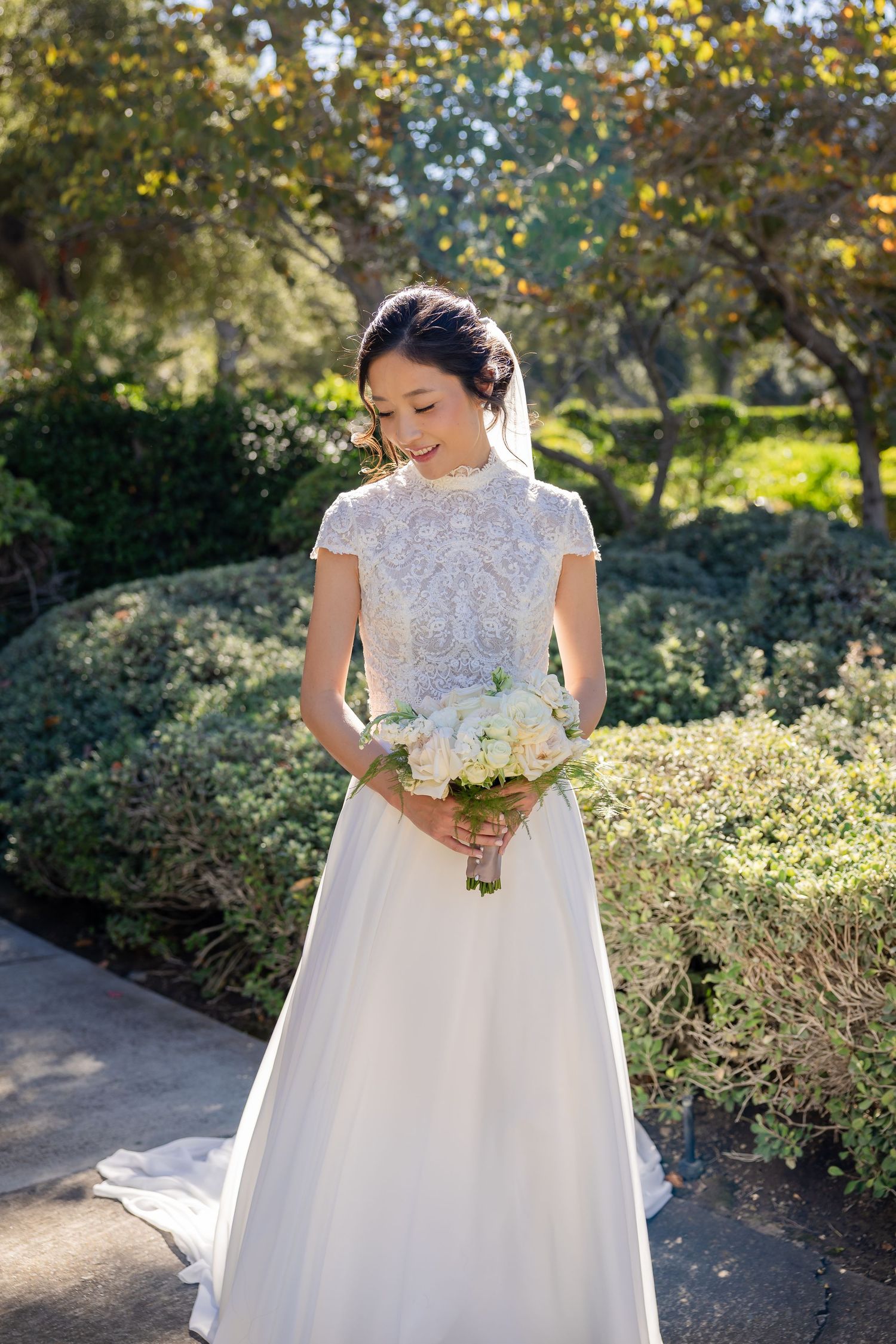 Elegant bride in lace high-neck wedding gown holds white bouquet while standing in a sunlit garden.