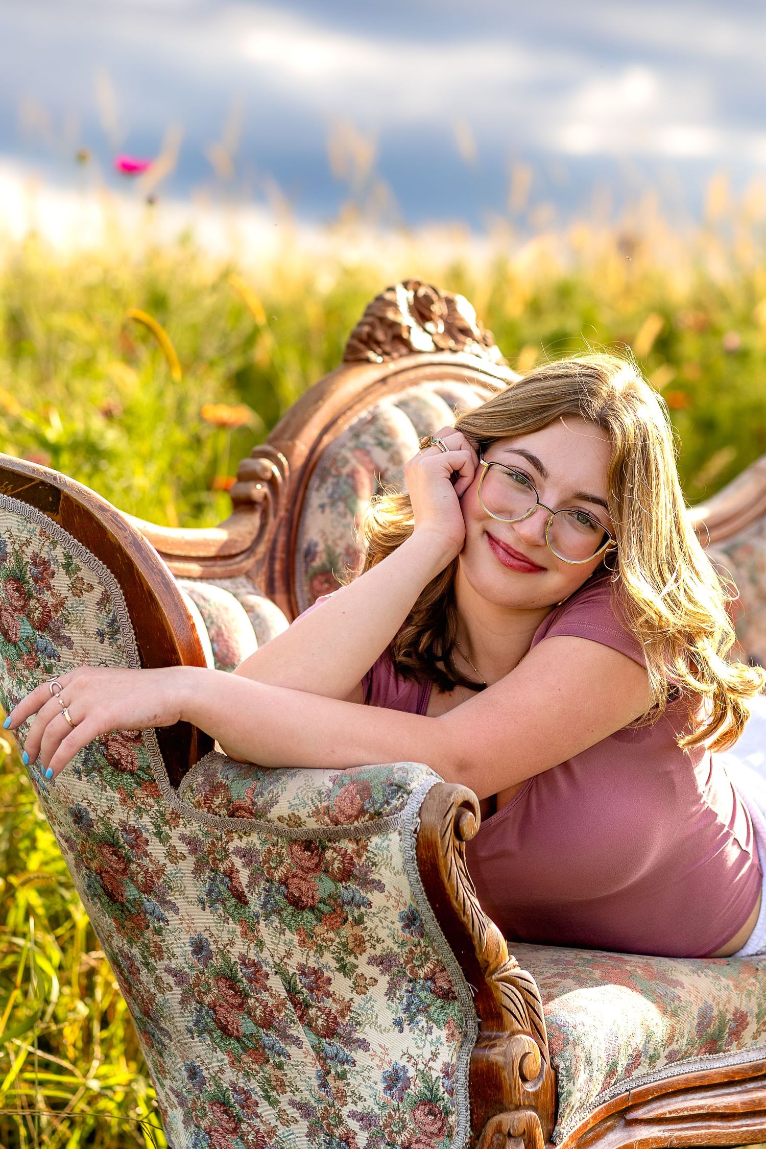 Smiling blonde teen with glasses reclining on ornate floral sofa in sunny wildflower field.