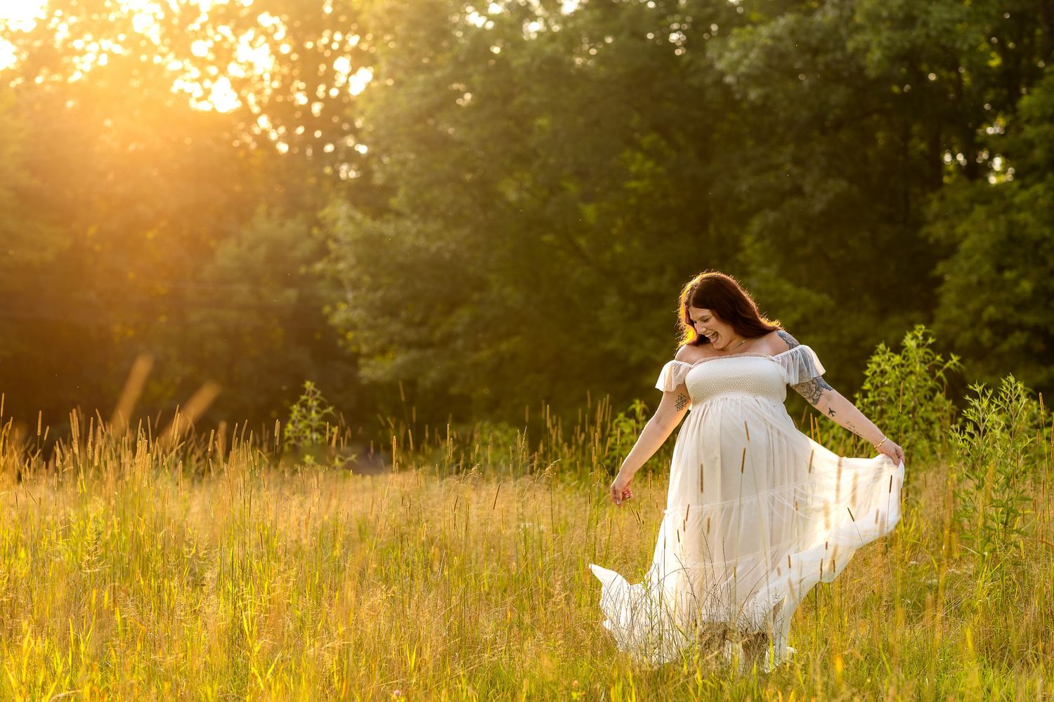 Pregnant woman in flowing white off-shoulder gown twirling in a sunlit green meadow at golden hour.