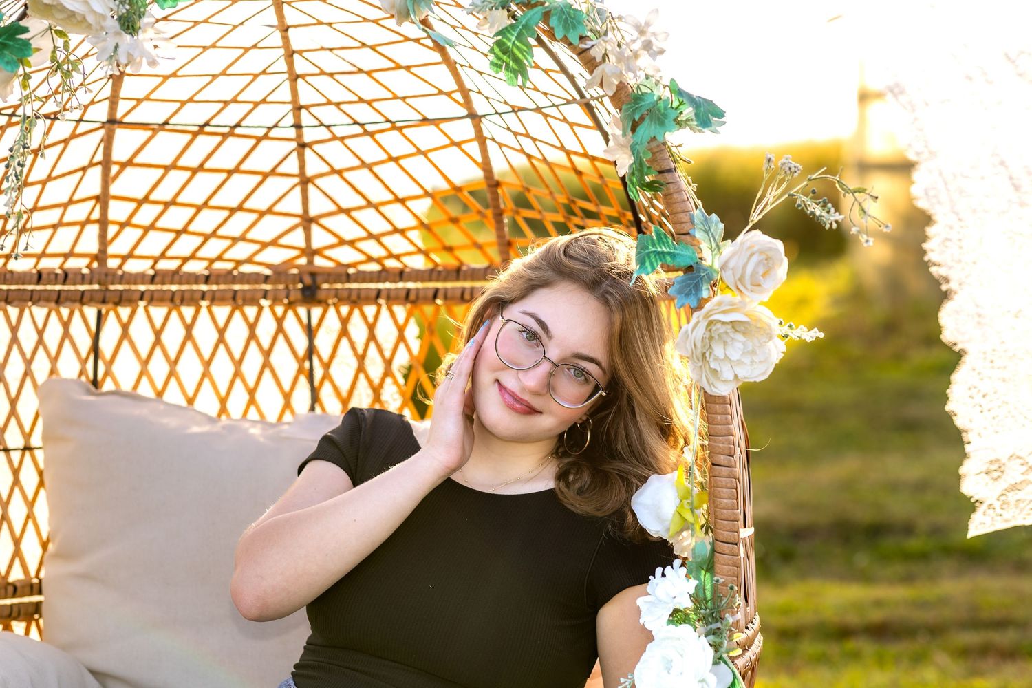 Smiling teenage girl with glasses relaxing in a decorative wicker swing chair surrounded by flowers outdoors.