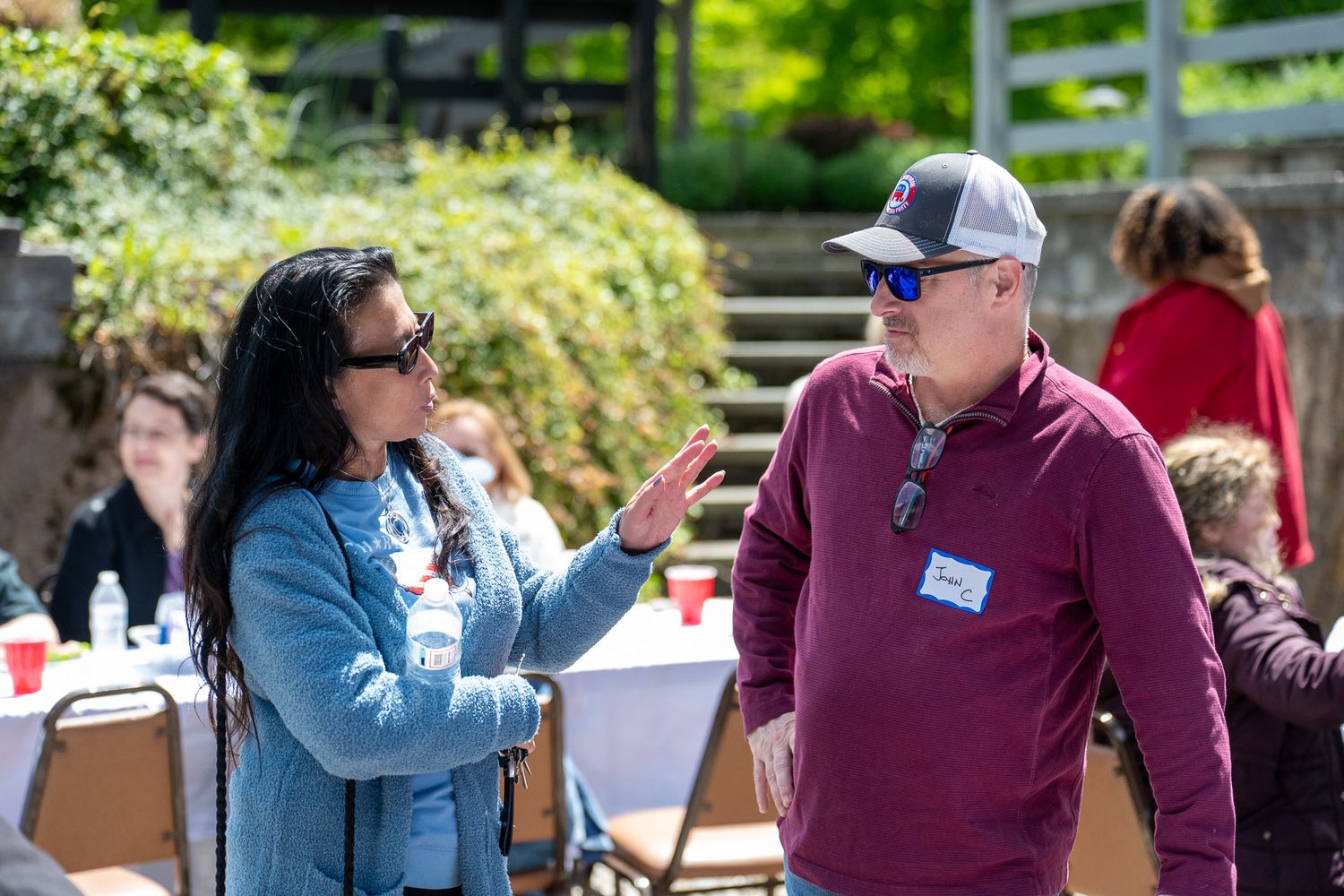 Two people in sunglasses engage in conversation at an outdoor gathering with tables and guests in the background.