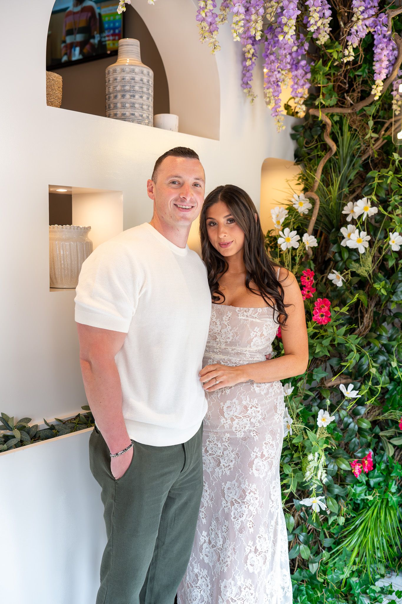 Smiling couple posing together in front of a lush floral wall with wisteria and colorful blooms at bridal shower.