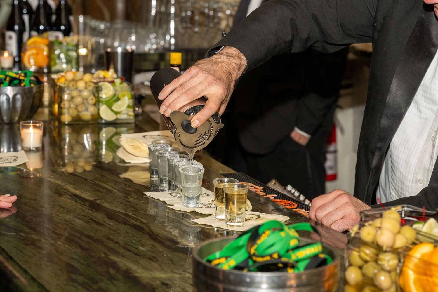 Bartender pouring shots into glasses lined up on a bar counter with garnishes and bottles in background.