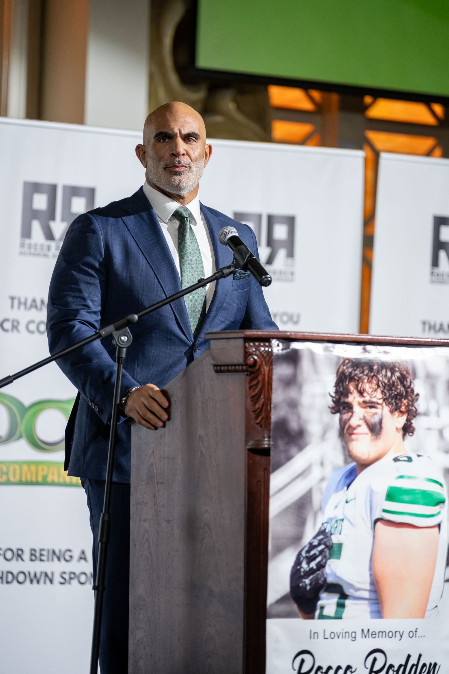 Man in blue suit speaking at podium during memorial event with green banner and tribute photo visible.