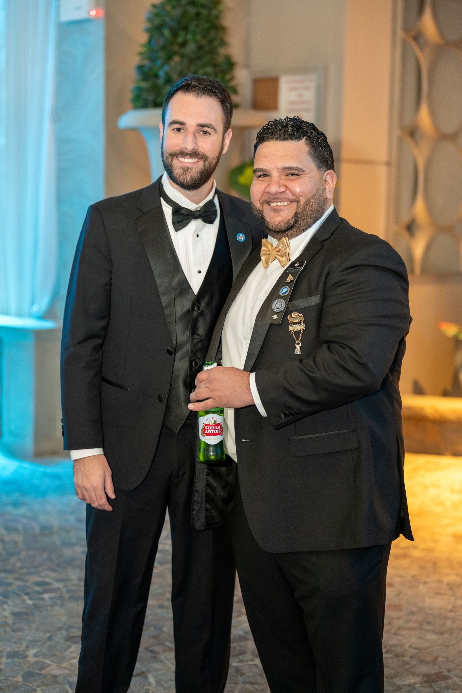 Two men in black tuxedos smile at a formal event, one holding a green beer bottle and wearing a gold bow tie.