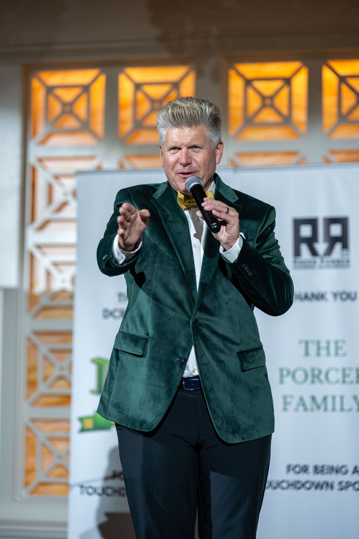 Man in green velvet blazer with gold bow tie speaks into microphone at formal fundraising event on stage.
