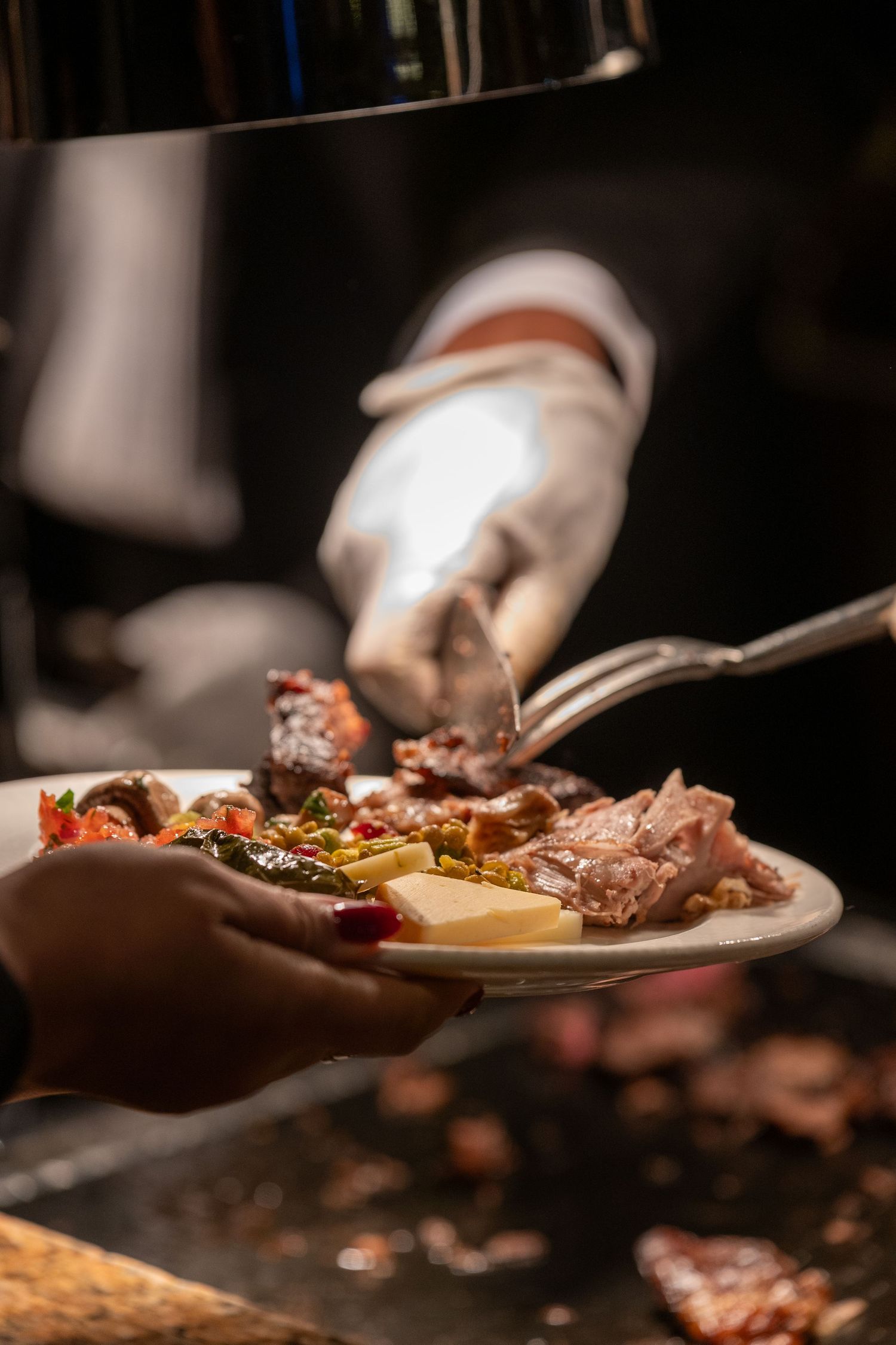Chef in white gloves serving grilled meat and vegetables onto a plate at a buffet restaurant.