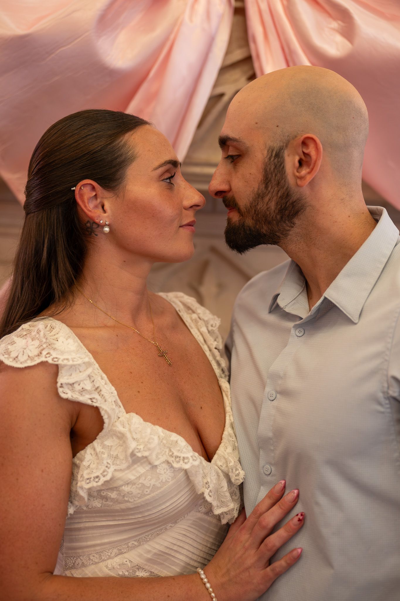 Romantic couple in wedding attire sharing an intimate moment under soft pink draped fabric backdrop.
