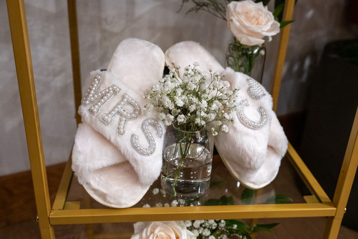 White fluffy MRS bridal slippers with pearl letters displayed on gold shelf with baby's breath and pink roses.