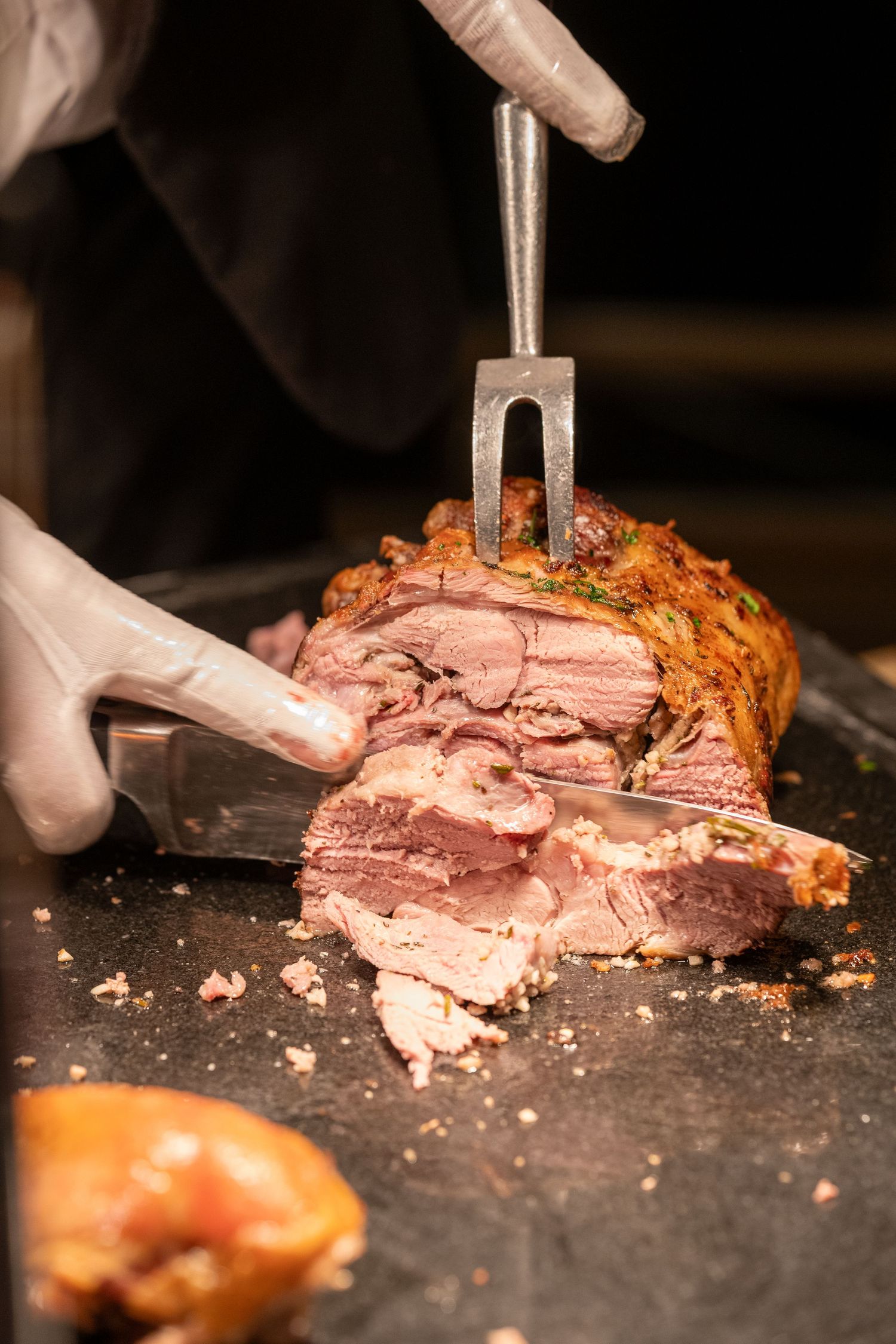 Chef carving a juicy roasted prime rib with a fork and knife on a dark cutting board.