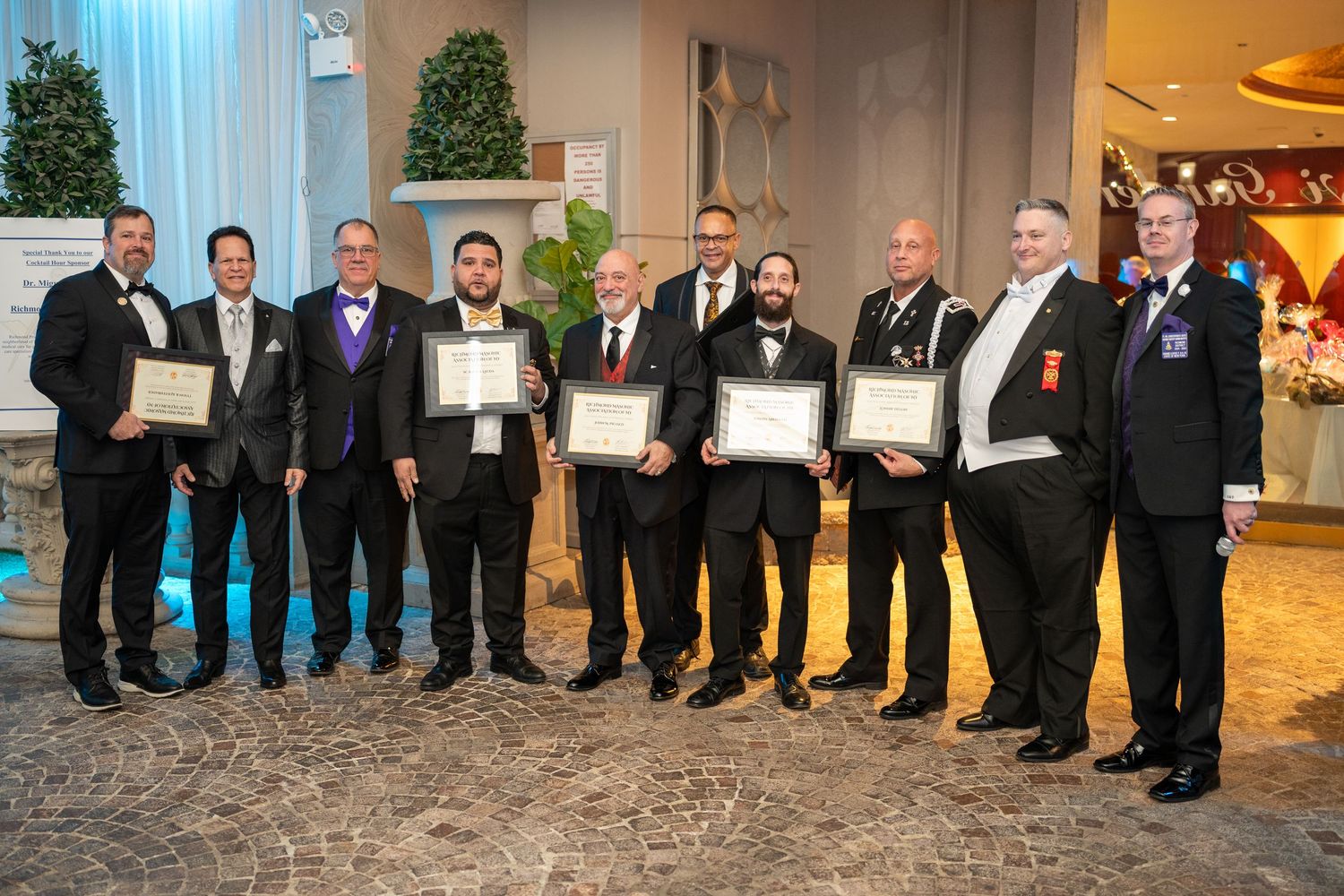 Group of men in formal attire posing with award certificates at an elegant banquet hall ceremony.