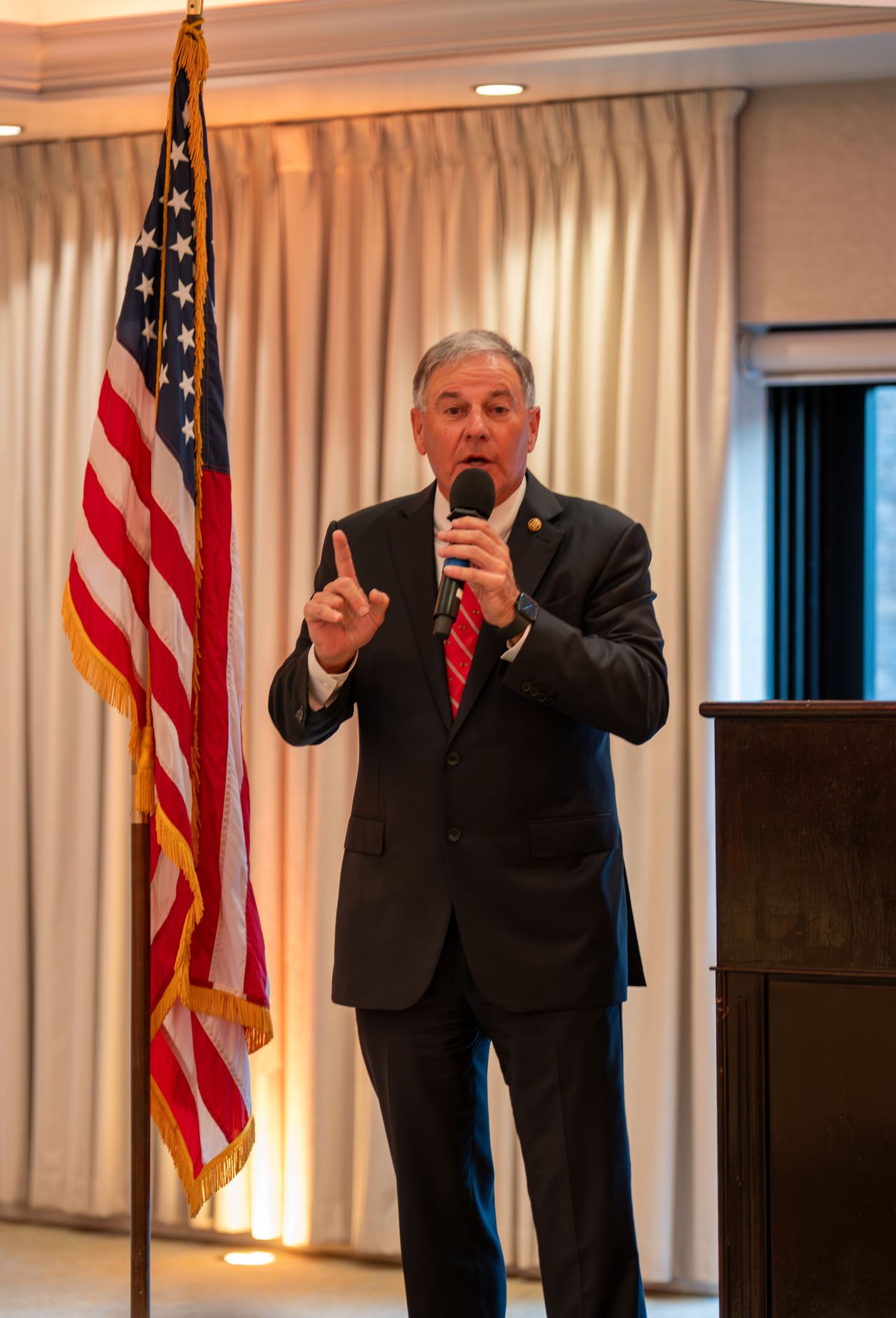 Man in dark suit speaking at podium with US flag, holding microphone and gesturing with finger raised.