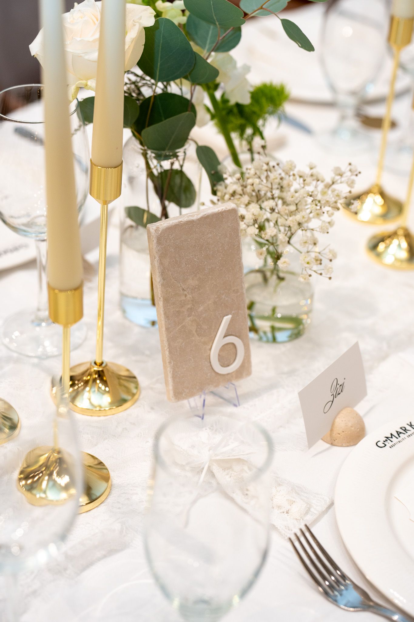 Wedding reception table detail with gold candlesticks, white florals, stone table number 6, and lace table runner.