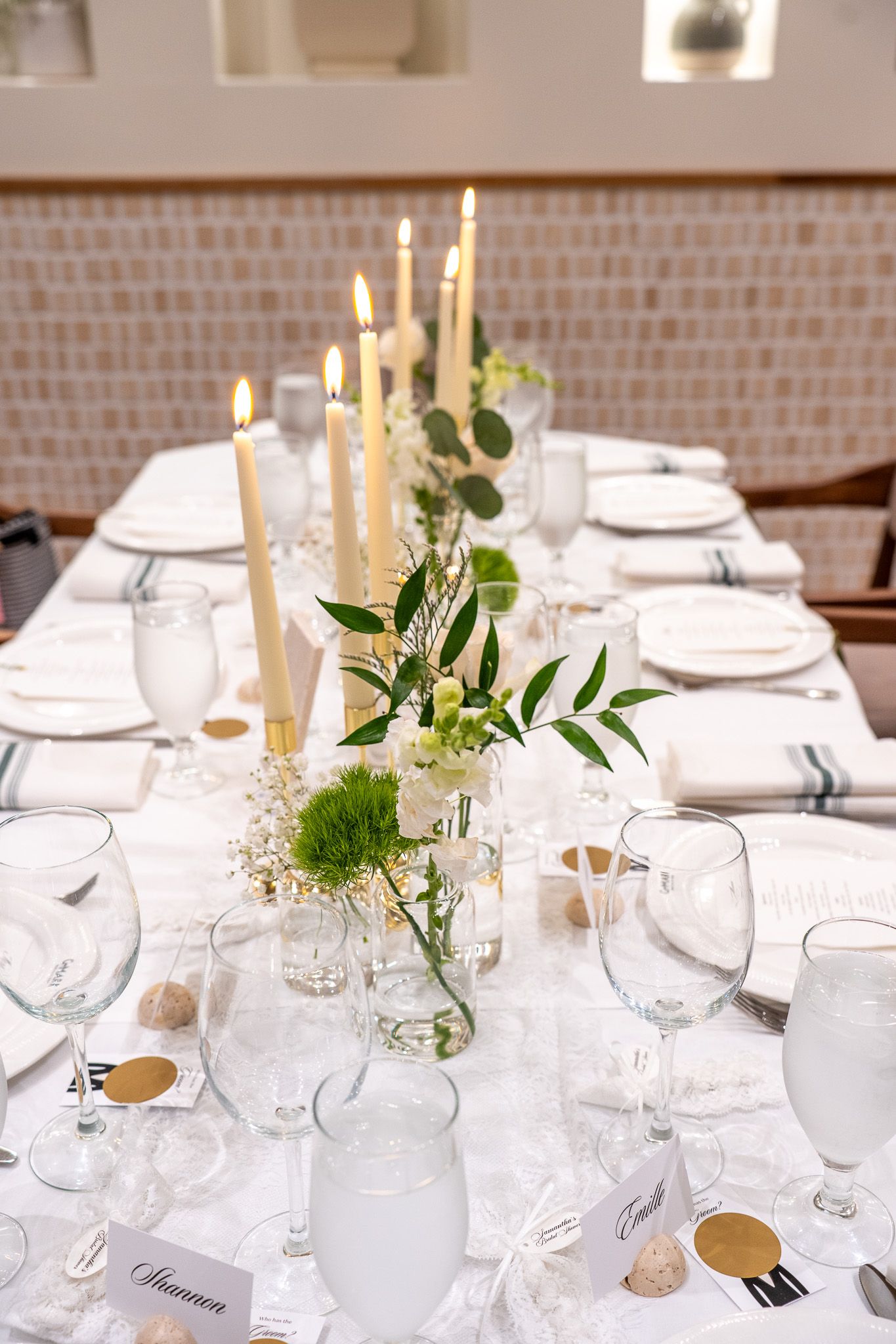 Elegant bridal shower table setting with white linens, lit taper candles, greenery centerpieces, and gold place cards.