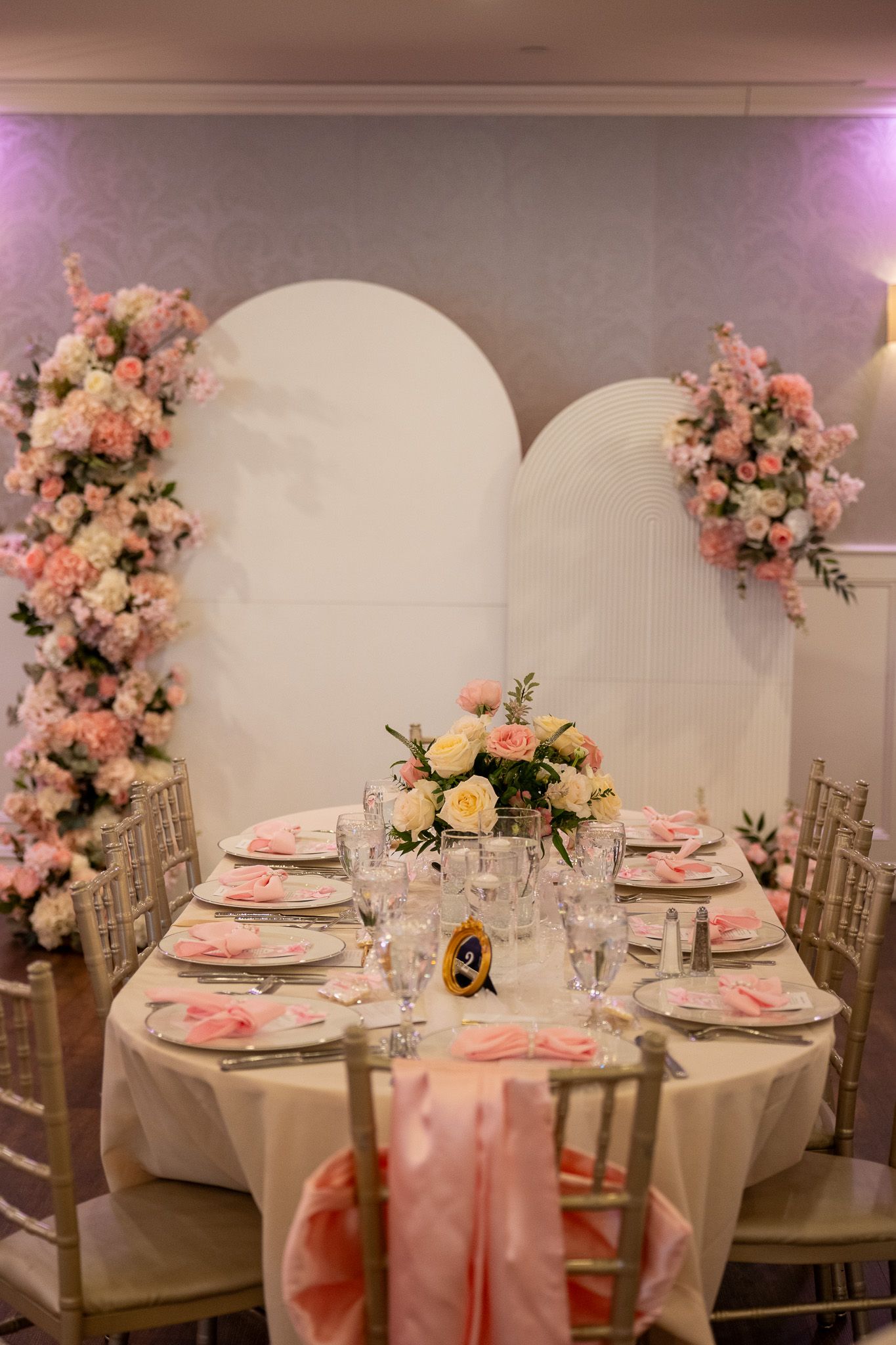 Elegant event table setting with pink floral arrangements, gold chiavari chairs, and arched white backdrop decor.