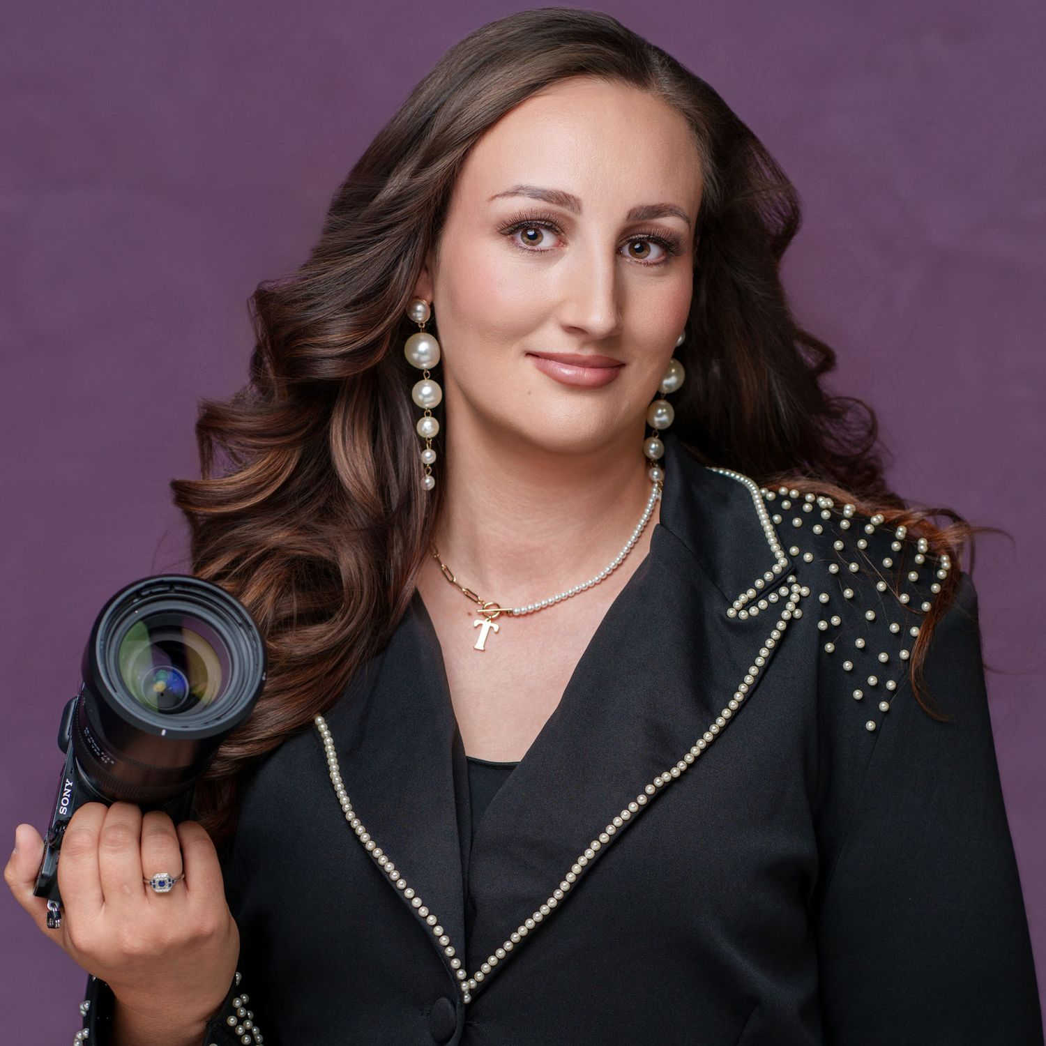 Professional headshot of person in black blazer with studded shoulders against purple backdrop.