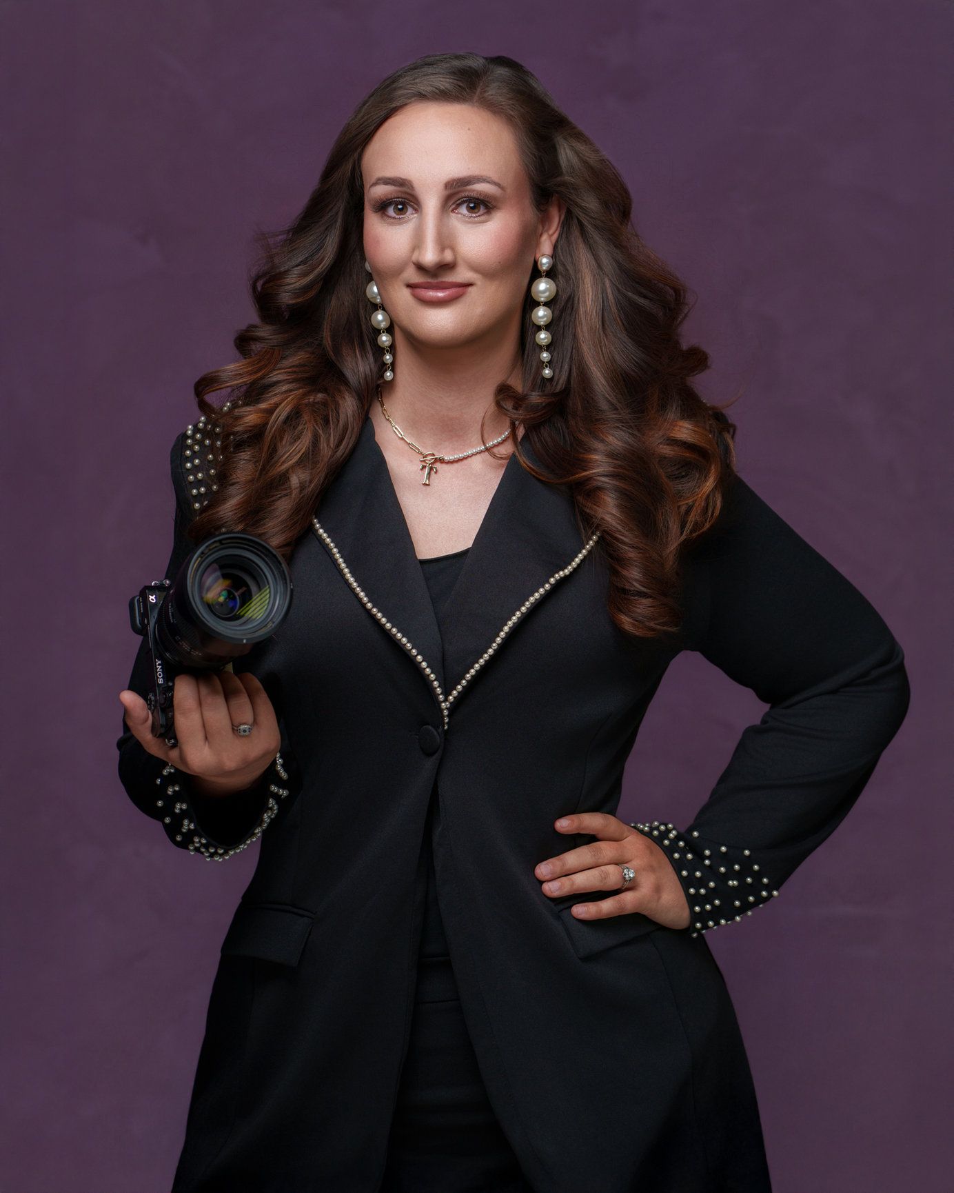 Professional portrait against purple backdrop featuring a person in black blazer holding camera.