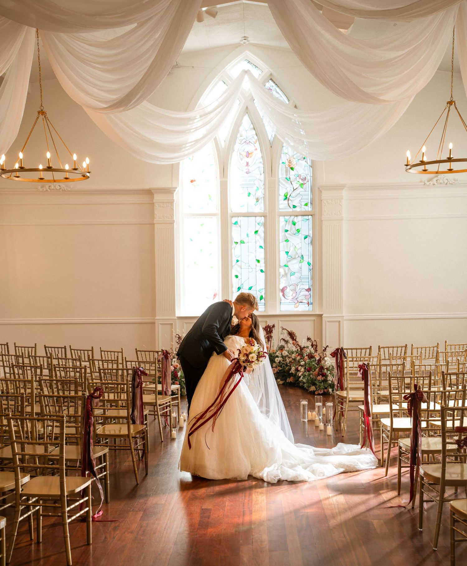 Wedding ceremony venue with draped white fabric, gold chairs, and arched windows.