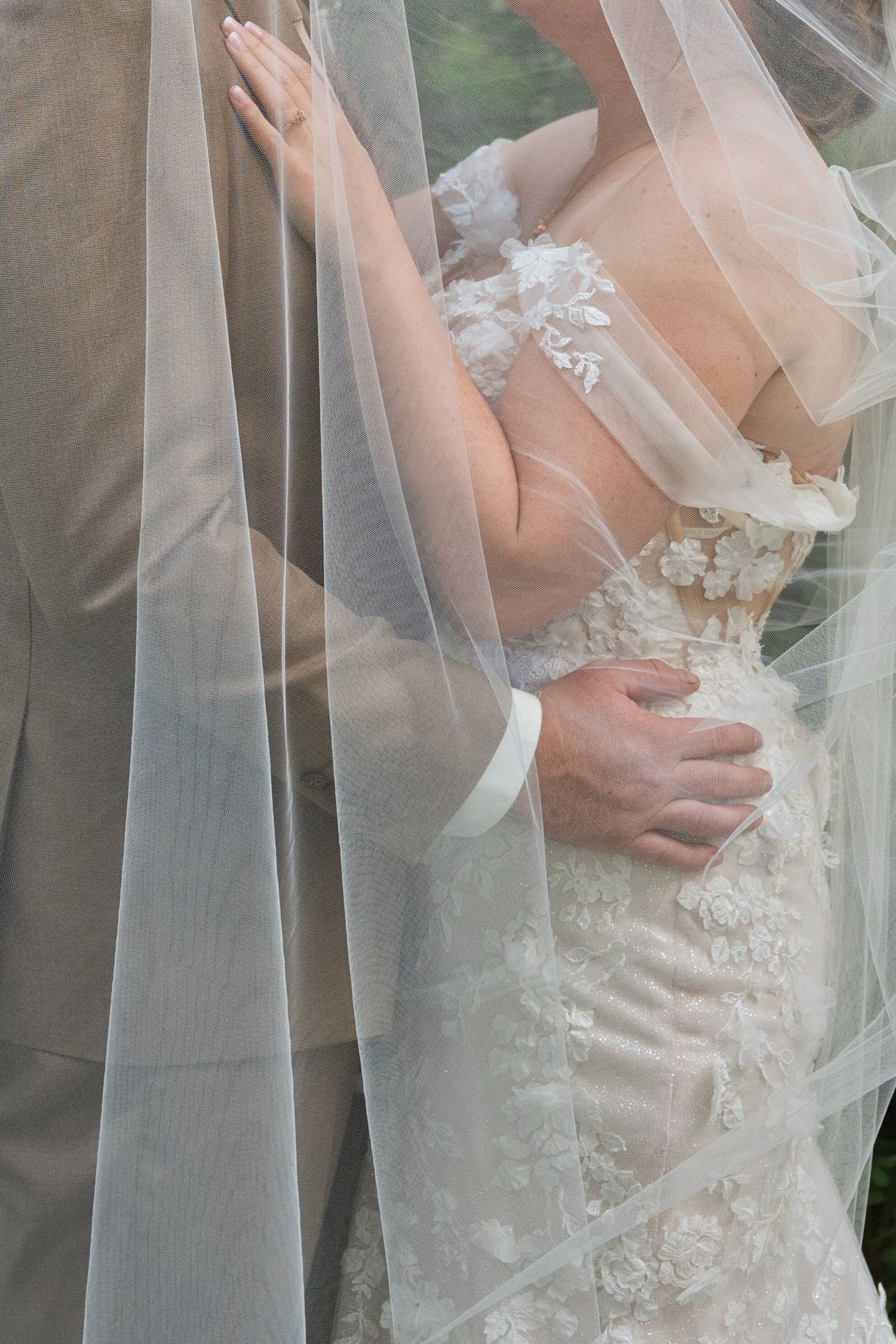 Dreamy photograph of a couple romantically embracing with a wedding veil draped over them, cropped to focus on their bodies.