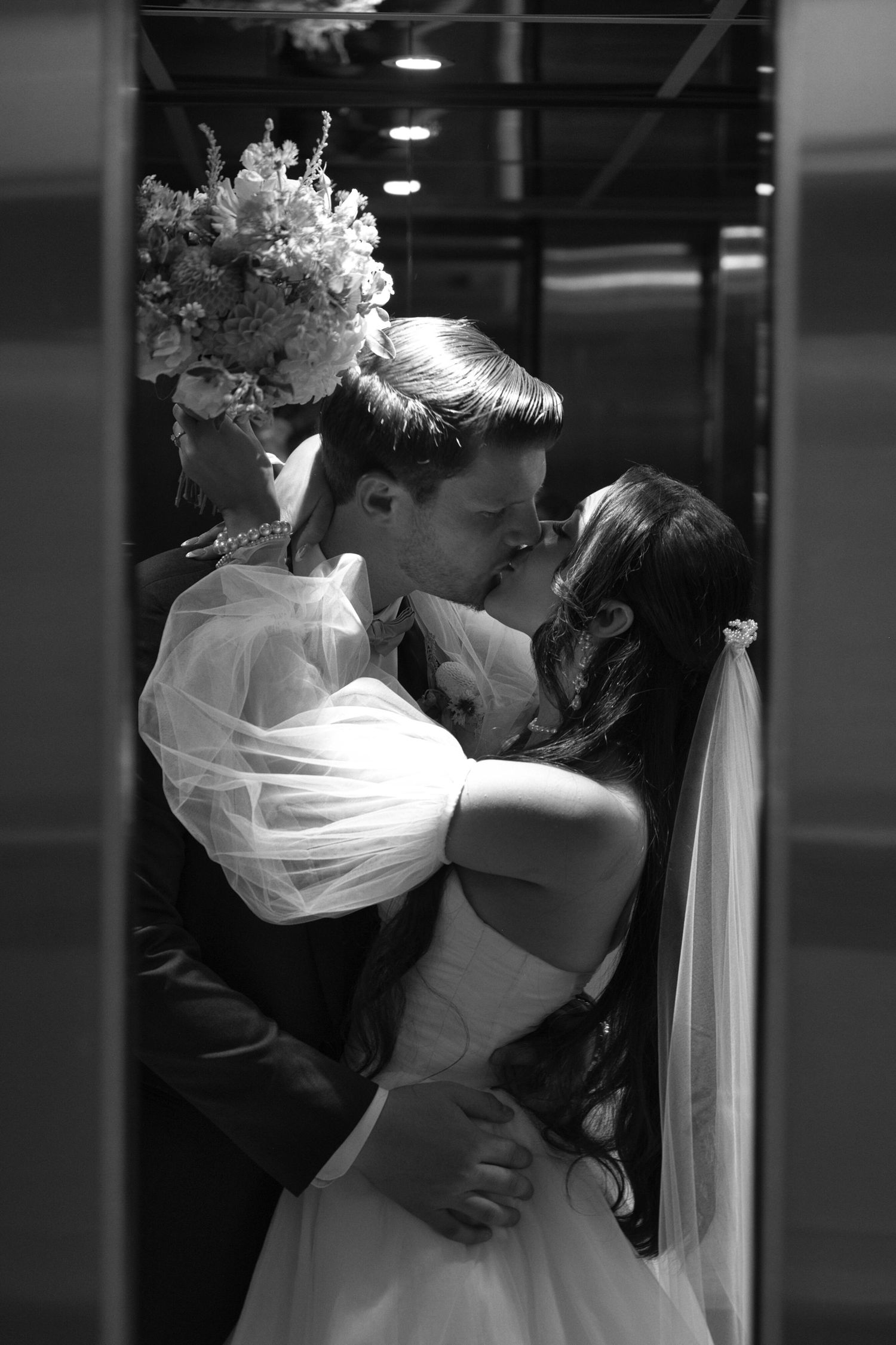 A romantic black and white portrait of a bride and groom kissing as the elevator doors are starting to close.