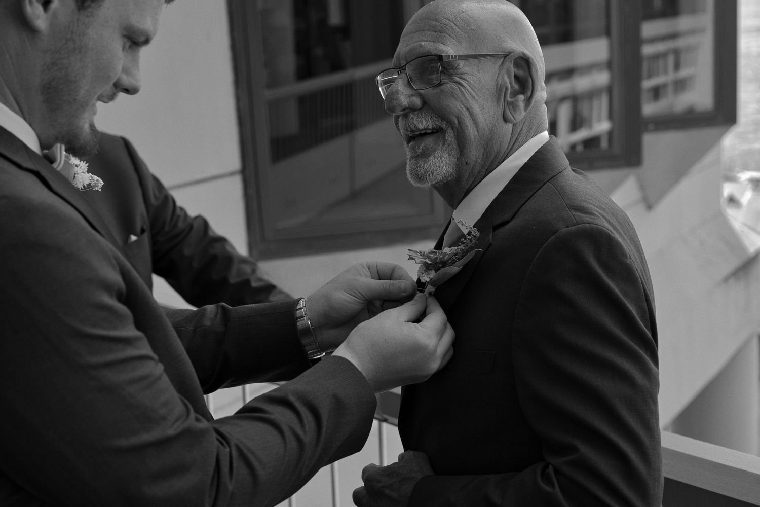 Black and white picture of the groom pinning the boutonniere to his grandfathers suit jacket while both men smile.