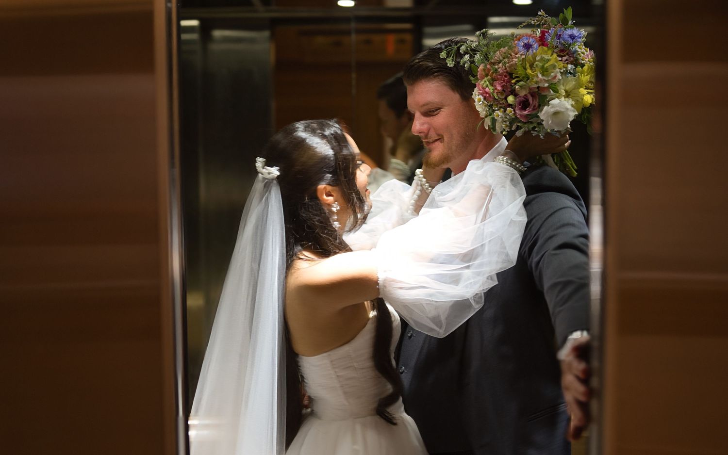 Groom holds an elevator door open as the bride reaches up to put her arms around his neck, ready to kiss him.