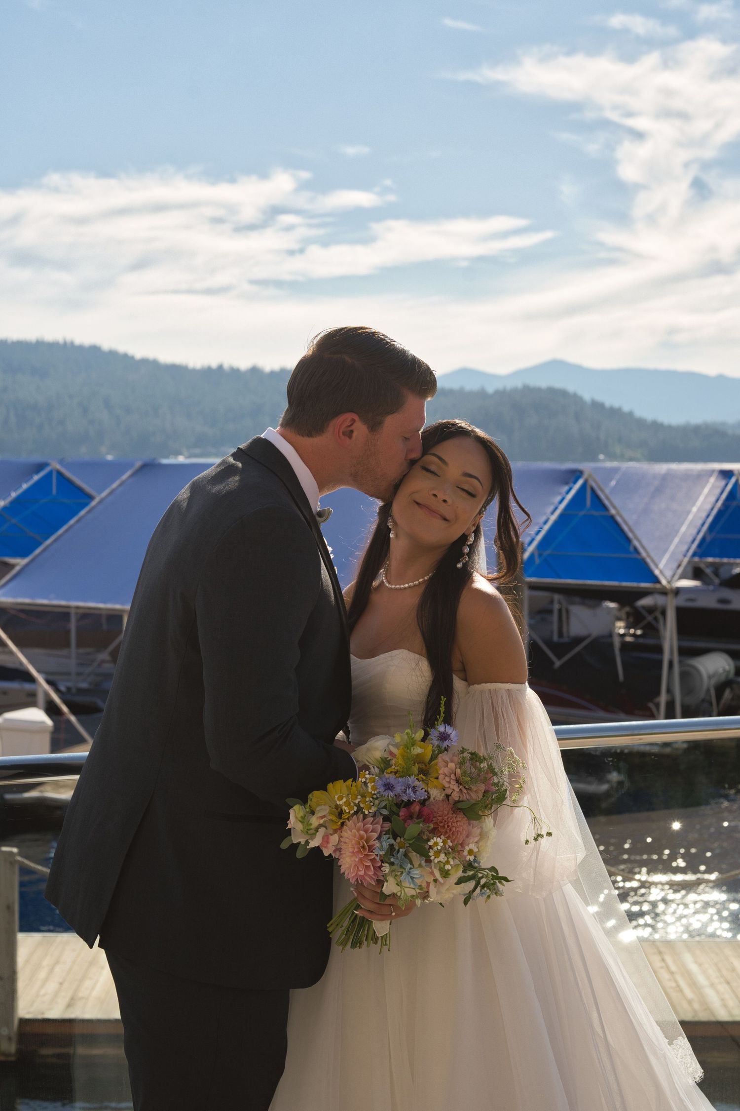 Groom kisses the bride's forehead as she smiles at the camera with her eyes closed as they stand outside.